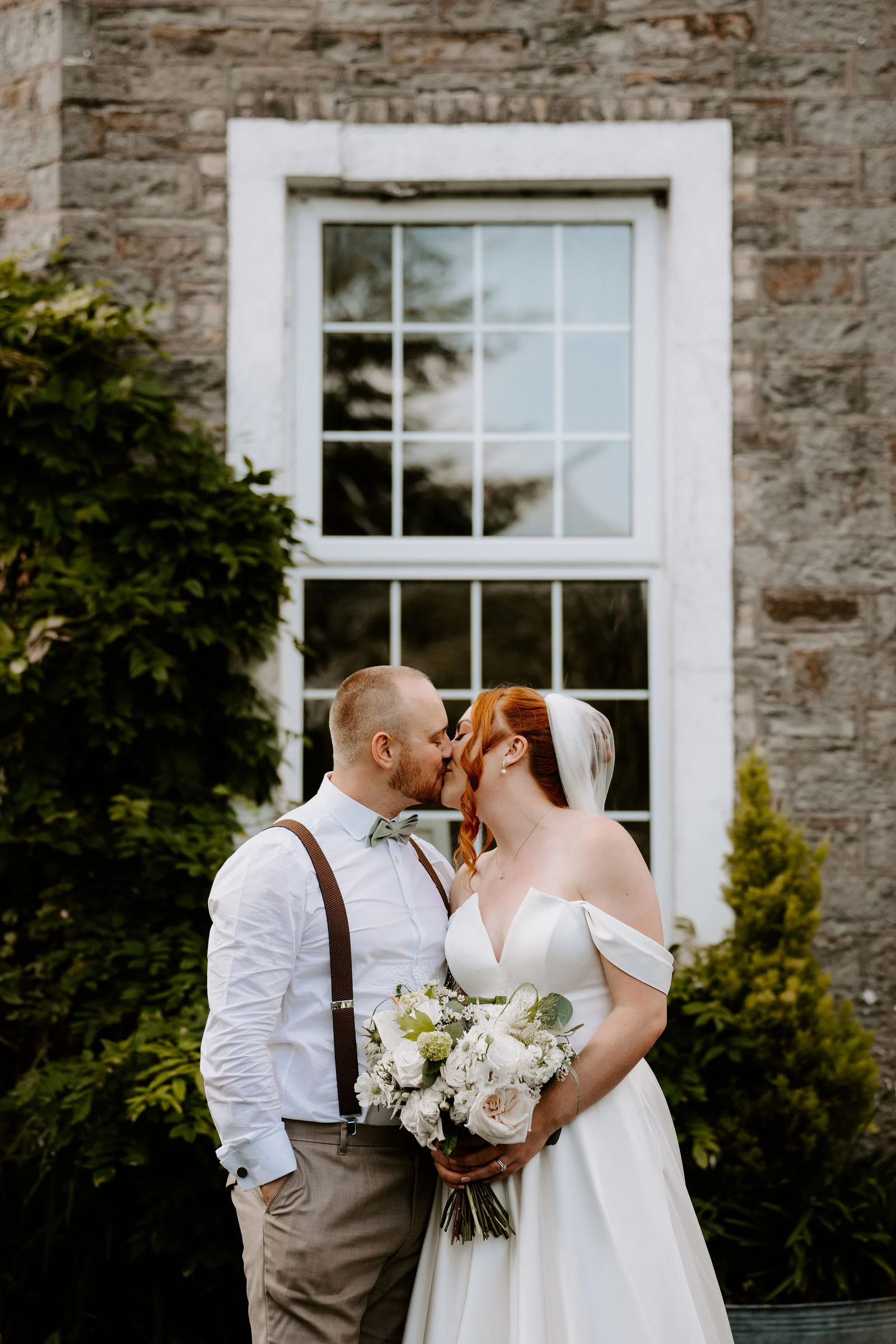 A groom in a white shirt and braces kisses his bride beneath a tall white-framed sash window in a stone wall, with a bouquet of white roses beside them