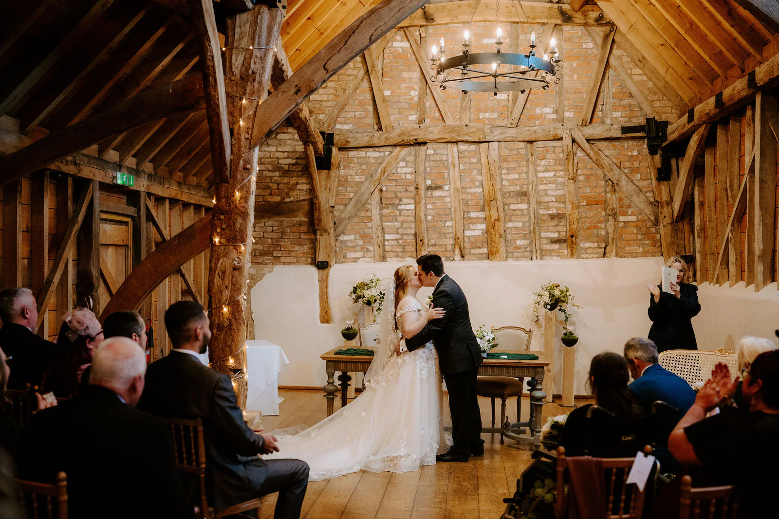 Georgia in a long veil and James kiss at the signing table in the brick and timber barn beneath an iron chandelier while guests watch