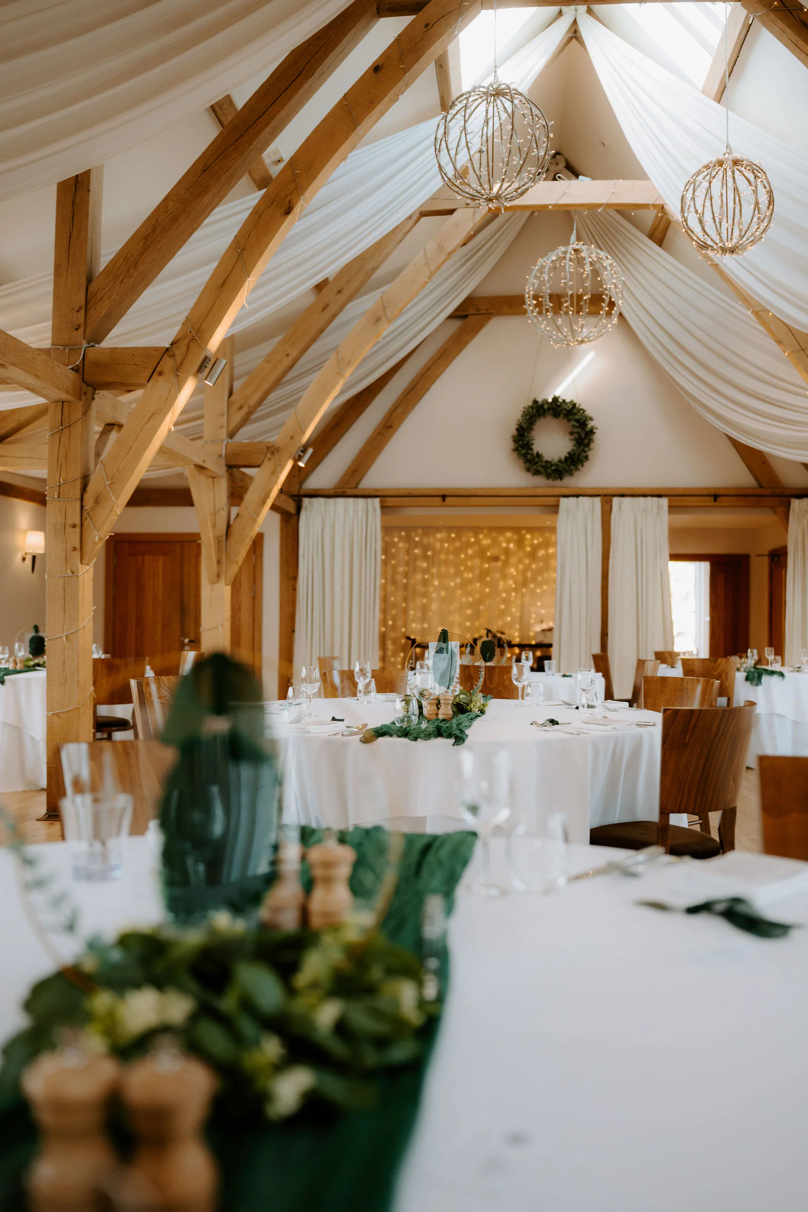 A table laid with white linen and dark green runners in a timber barn with a draped white ceiling and hanging round lights at Bassmead Manor Barns