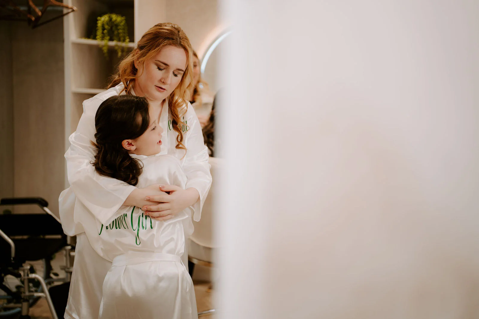 Georgia in a white robe holds a young flower girl in a matching robe in a bright room before the ceremony