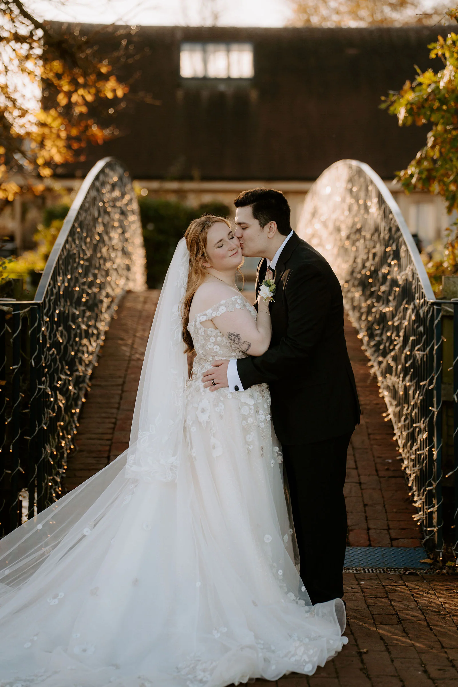 James kisses Georgia on the cheek on a brick path between the arches of a fairy-lit footbridge, her long veil and tulle gown around them