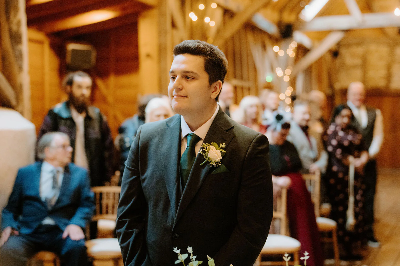 James in a dark green suit waits at the front of the timber barn as guests look on behind him