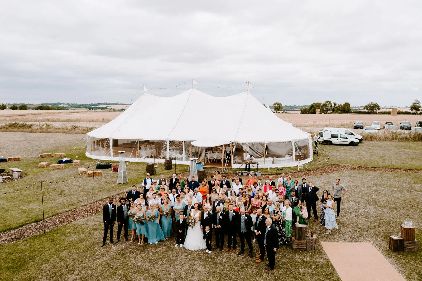 From above, wedding guests in bright colours gather on the lawn in front of a white sailcloth marquee in open farmland
