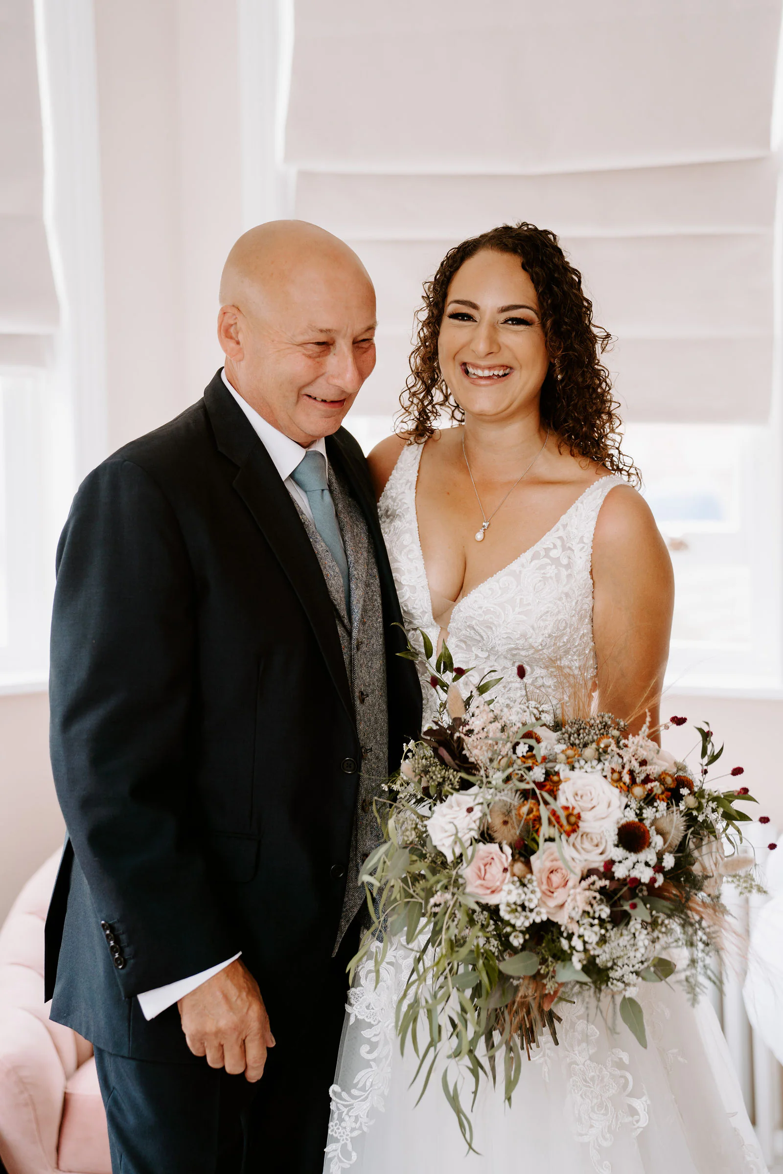 Kelly in a lace gown holds a bouquet of blush and dried flowers beside her father in a navy suit as they laugh together in a bright room