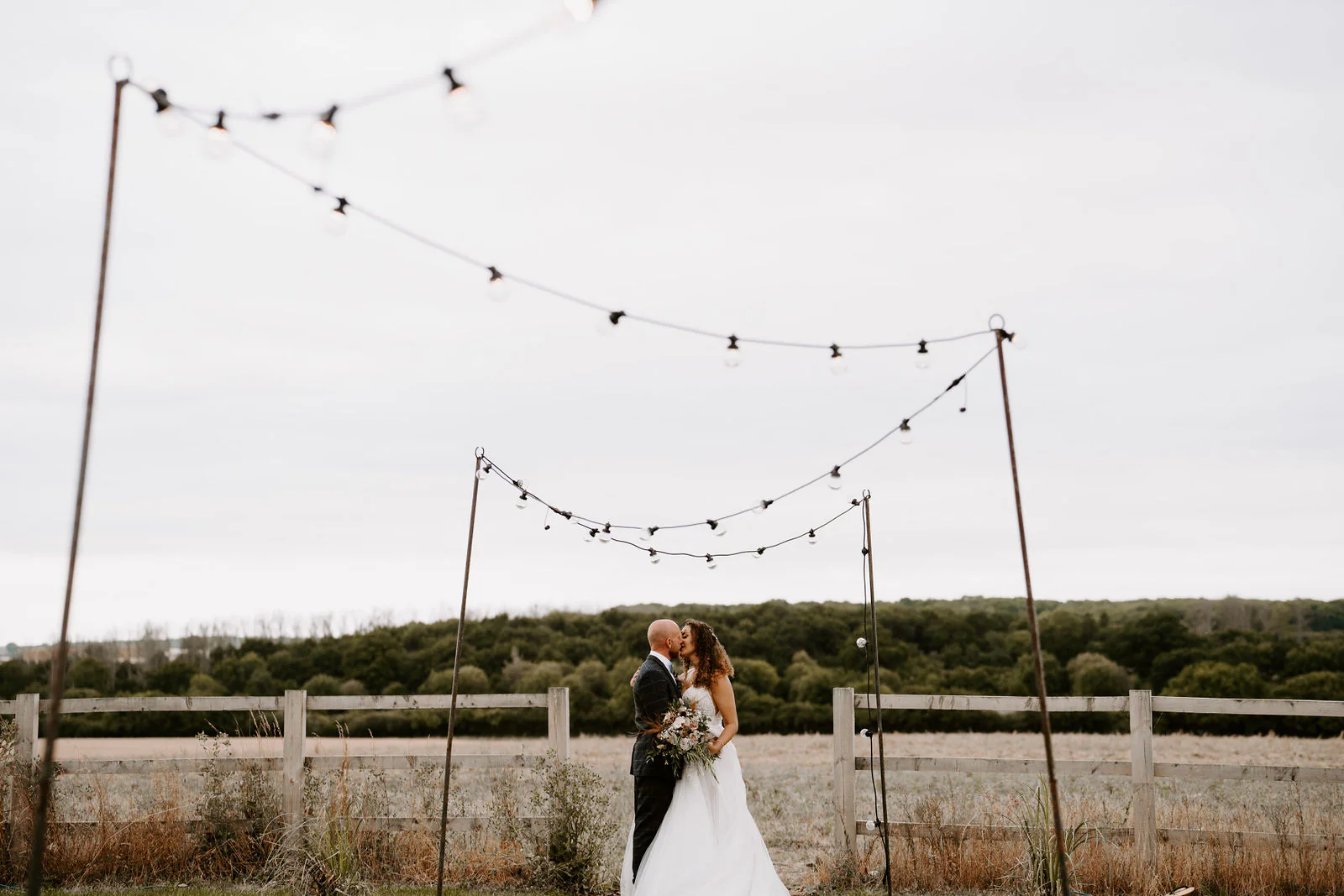 Josh and Kelly kiss beneath strings of festoon lights hung from tall posts, with a wooden fence and a dark line of woodland behind them