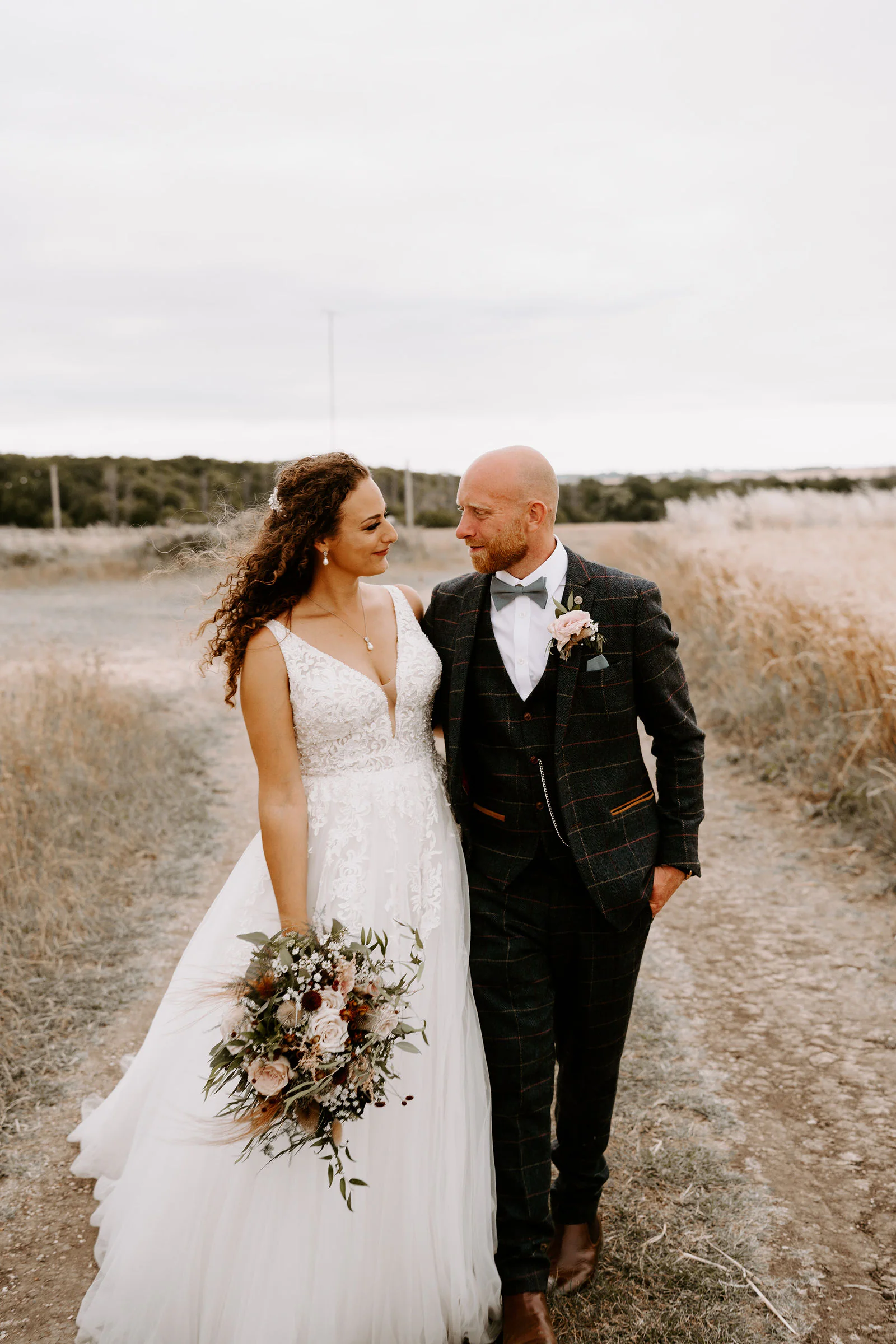 Kelly and Josh walk side by side along a grass track between fields, Kelly holding a bouquet of blush and dried flowers