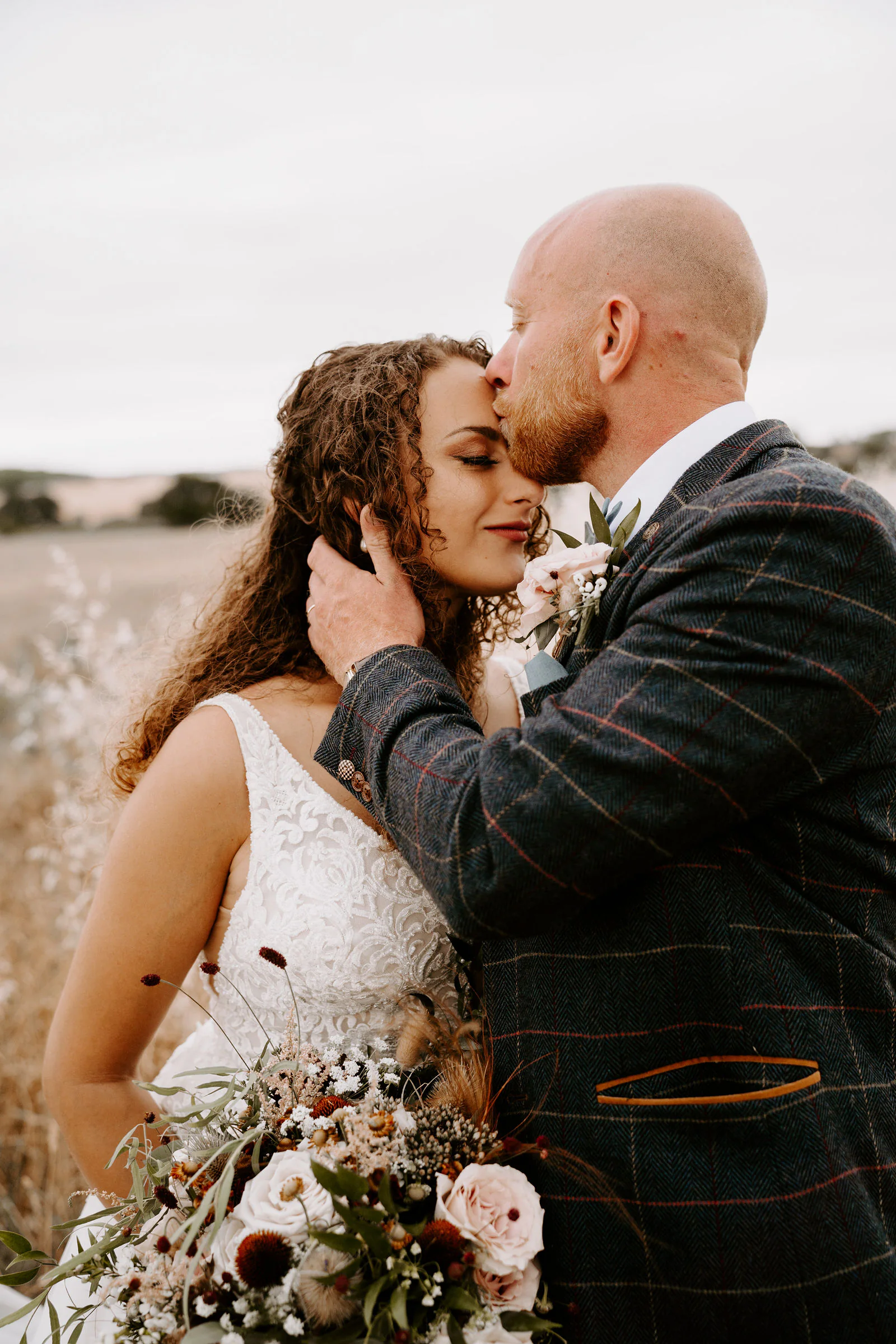 Josh kisses Kelly on the forehead as she touches his face, a bouquet of blush roses and dried flowers in front of them in a field of pale grass