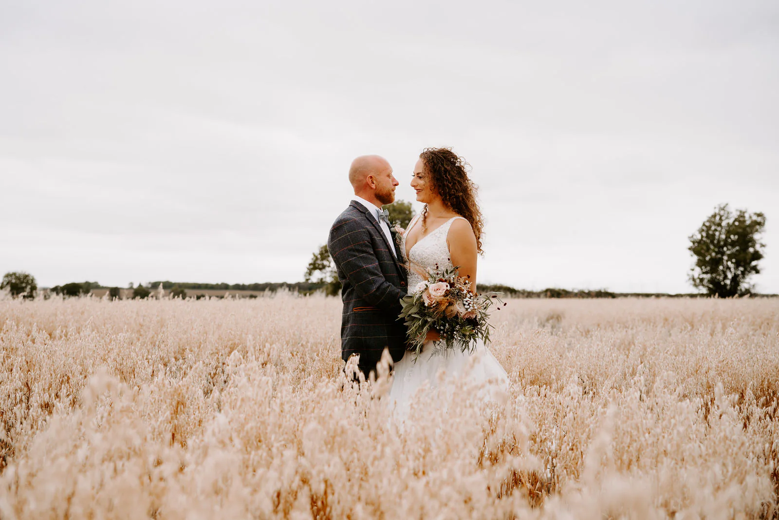 Kelly and Josh hold each other in a field of pale golden oats under a grey sky