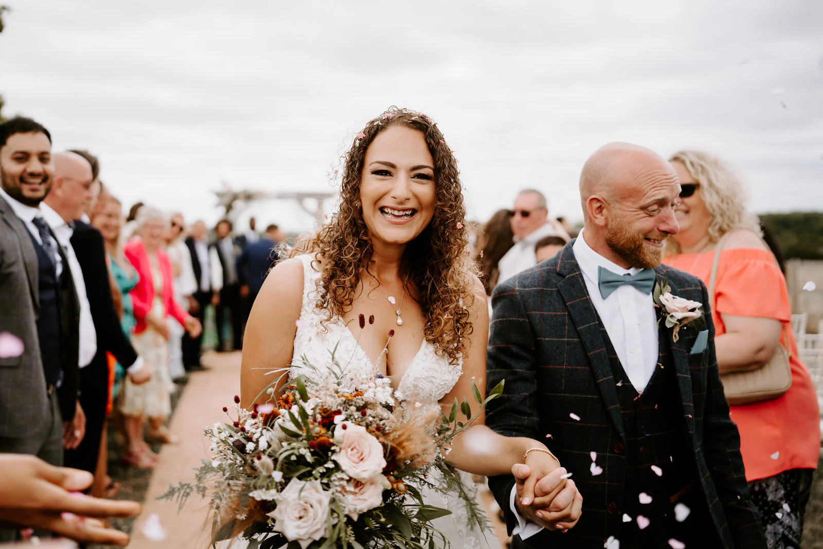 Kelly and Josh walk hand in hand through thrown petals between rows of guests along a path