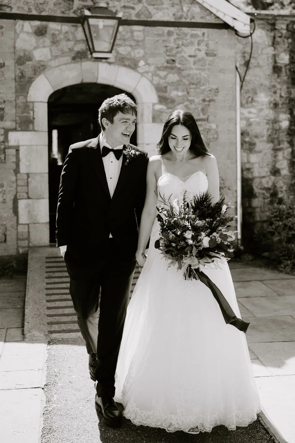 Amy and Dan walking hand-in-hand beneath the historic stone archway at Pencoed House Estate in Cardiff