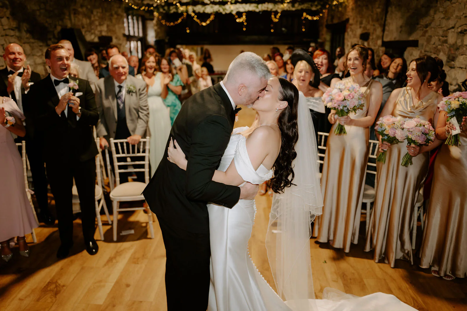 The couple's first kiss as a married couple at the end of the aisle in a stone-walled hall at Pencoed House Estate, guests applauding and bridesmaids in champagne satin holding pastel bouquets