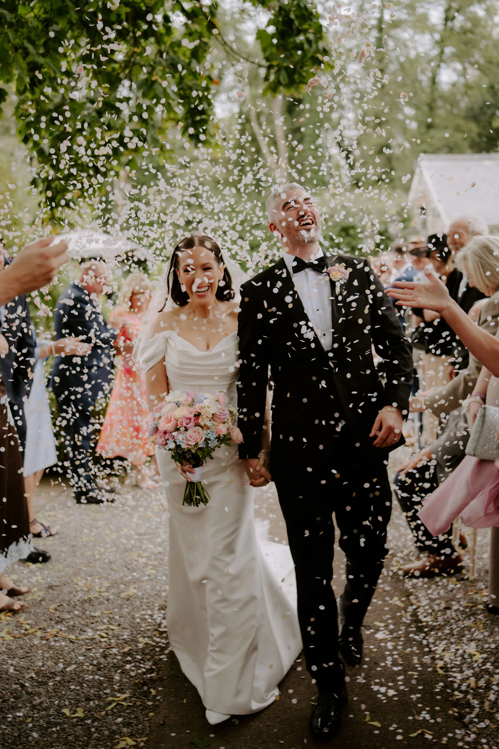 Faye and her groom laughing and holding hands as guests throw confetti on a tree-lined path at Pencoed House Estate