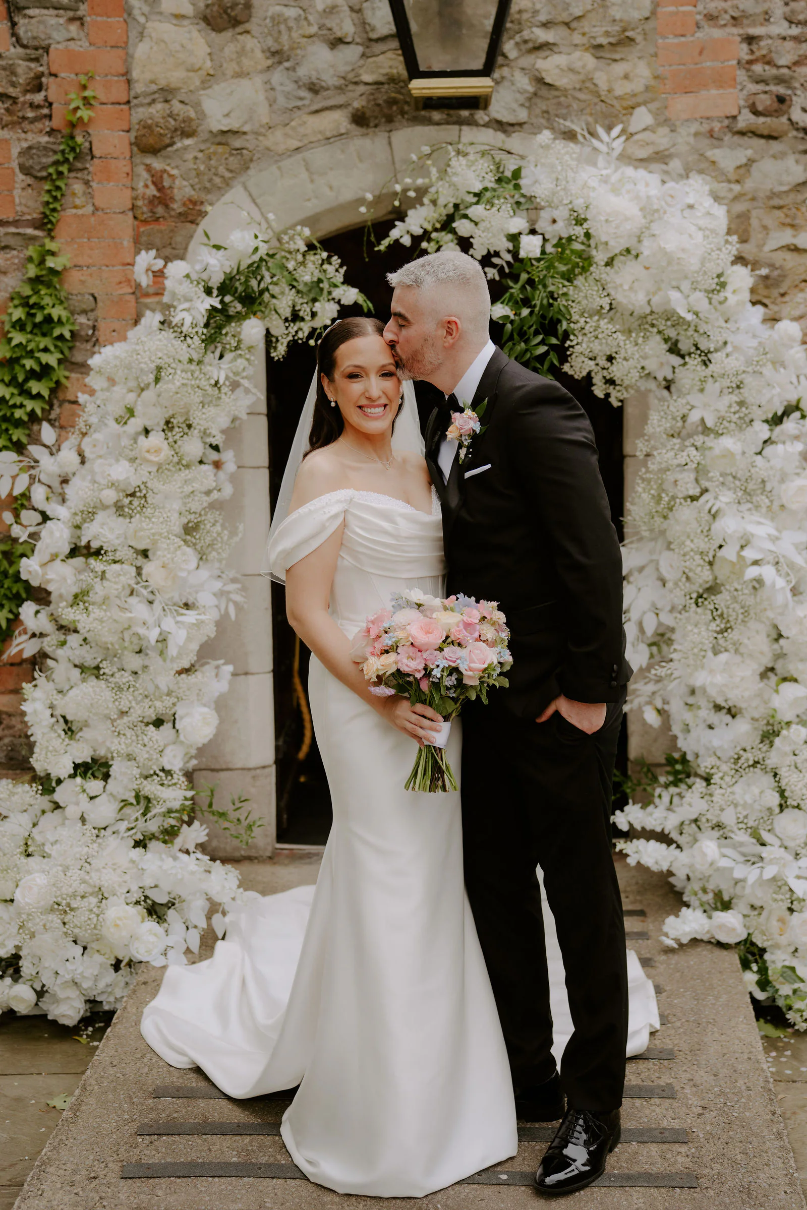 Faye and her groom beneath a stone archway dressed in white blossom at Pencoed House Estate, he kisses her cheek as she smiles and holds a pastel bouquet