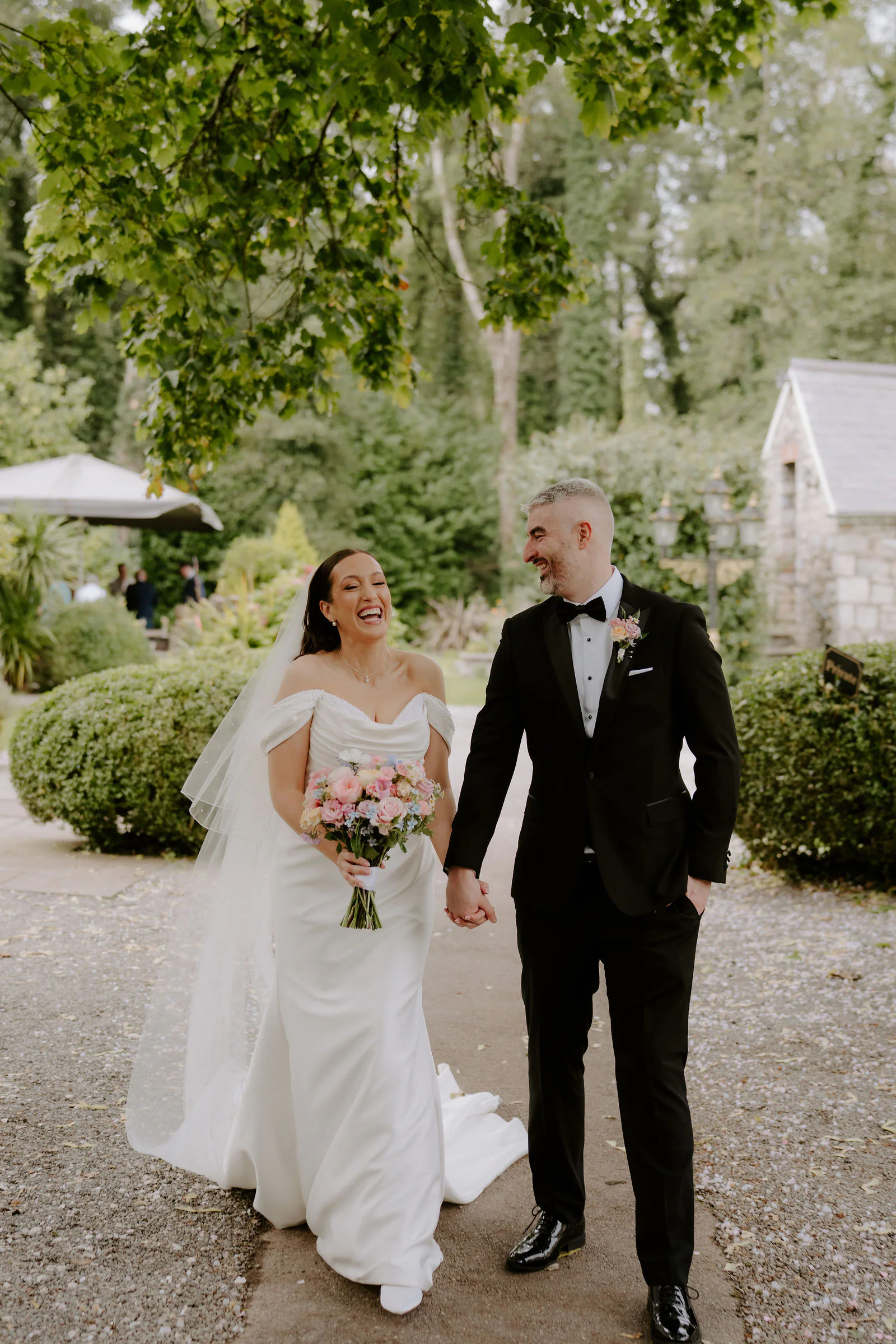The couple walking hand in hand along a gravel path at Pencoed House Estate, the bride laughing in an off-the-shoulder gown and veil with clipped hedges and a stone outbuilding behind them