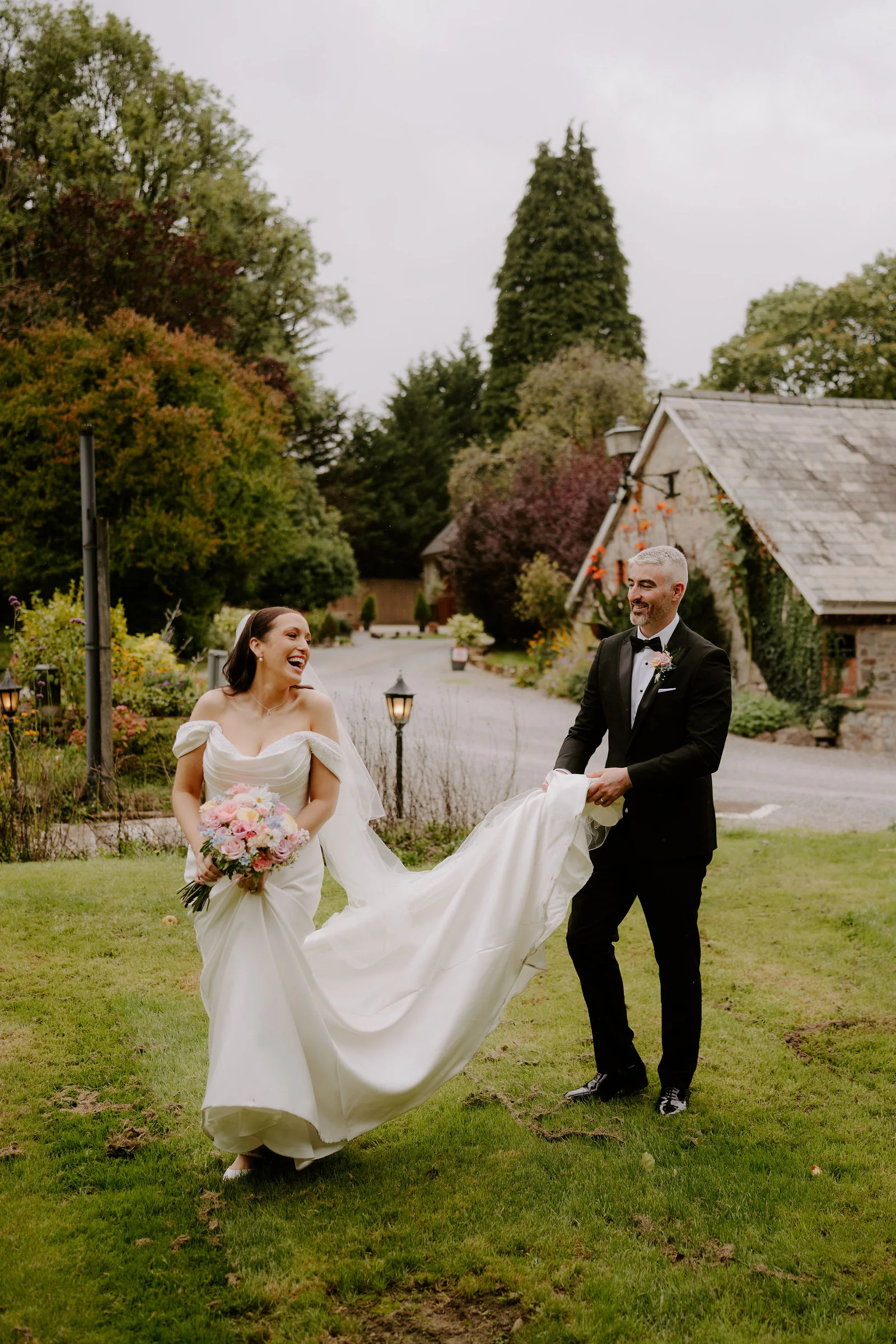 The bride laughing as she gathers her train across the lawn at Pencoed House Estate while the groom holds the end of it, with lamp posts, a tall conifer and a stone cottage behind them