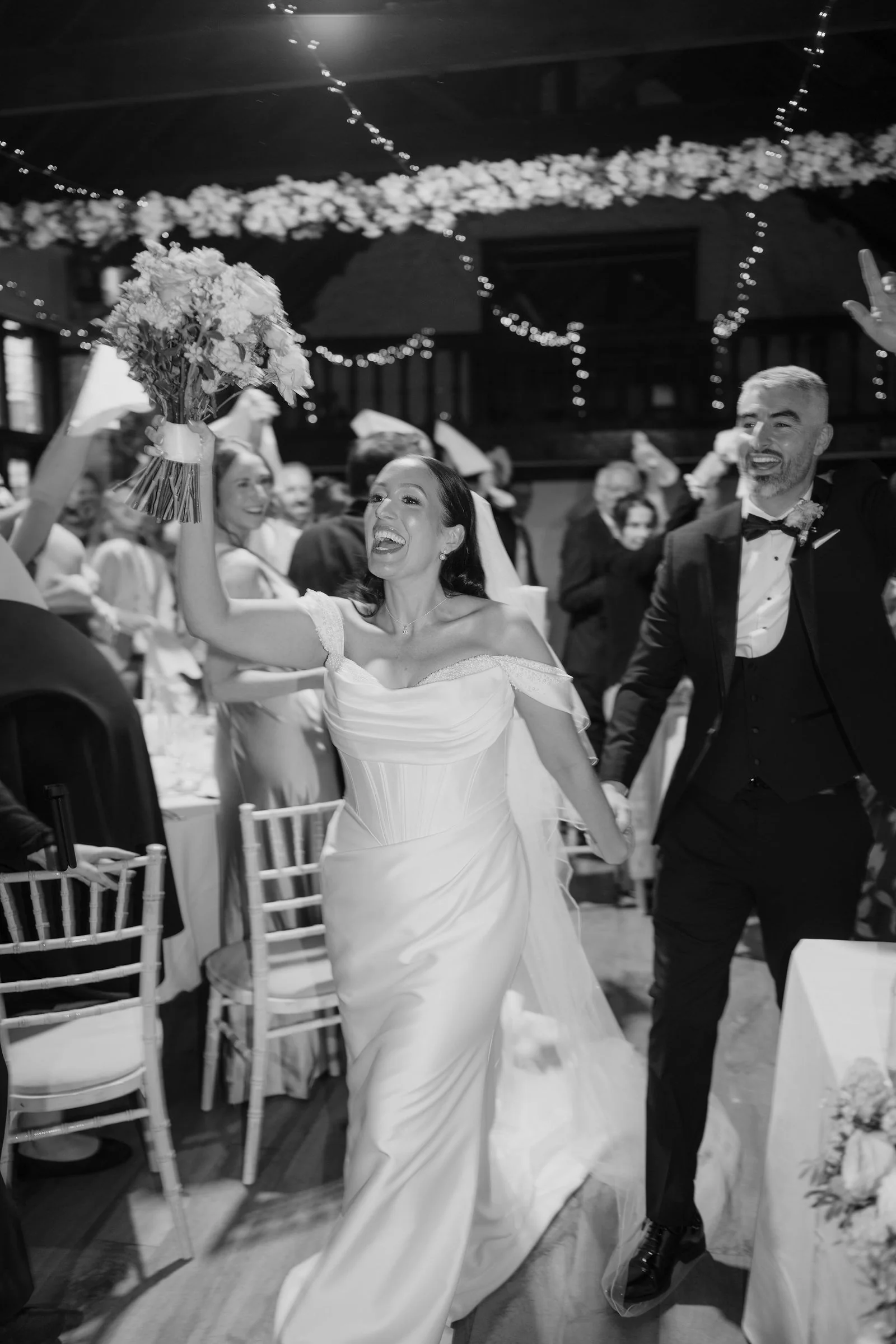 In black and white, the bride raises her bouquet and cheers as she and the groom walk hand in hand into the hall beneath hanging flowers and fairy lights, guests waving napkins around them