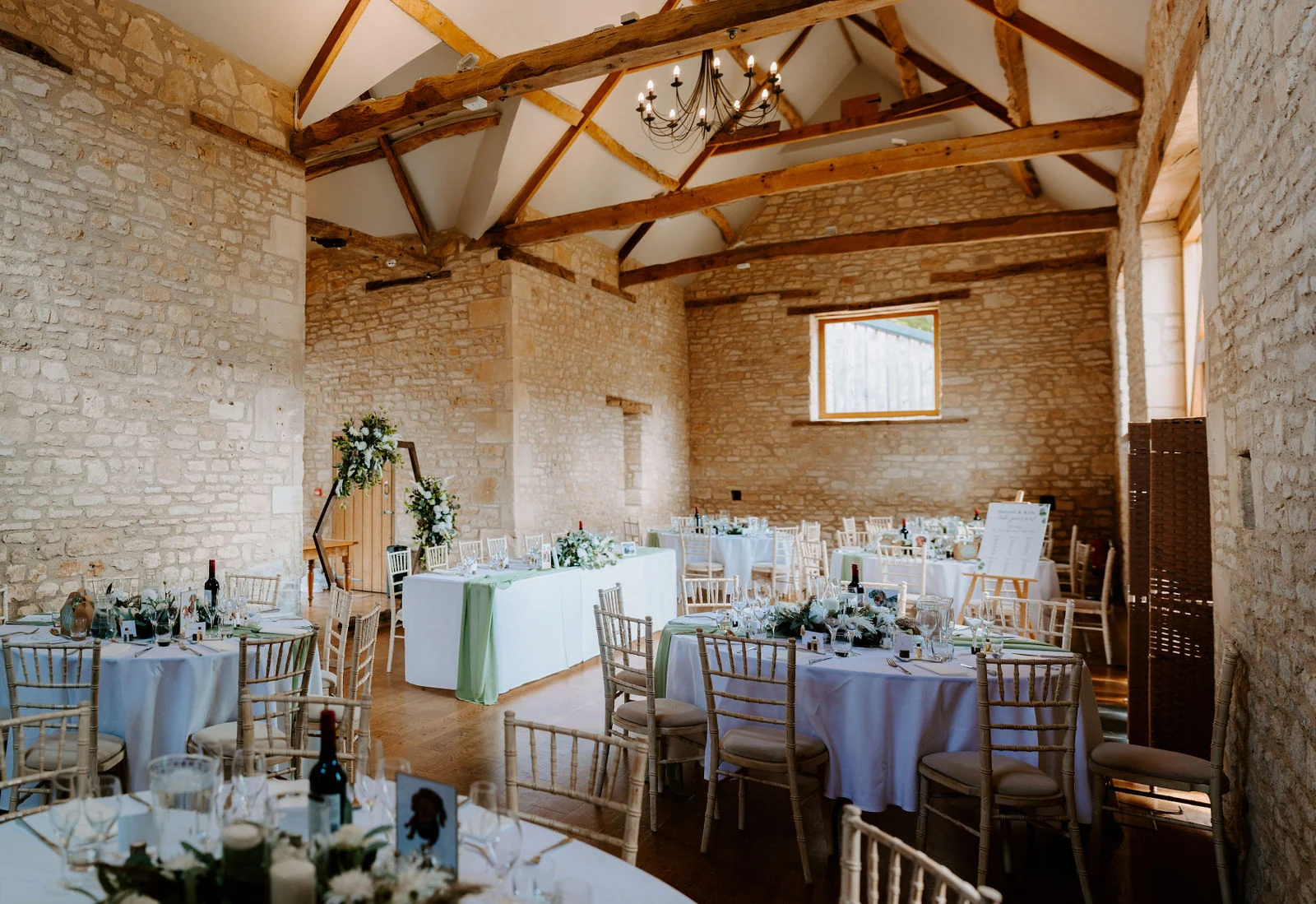 The inside of a stone barn with exposed oak roof beams and an iron chandelier, set with round tables in white linen, green table runners and pale wooden chairs, and a hexagonal floral arch at the far end.
