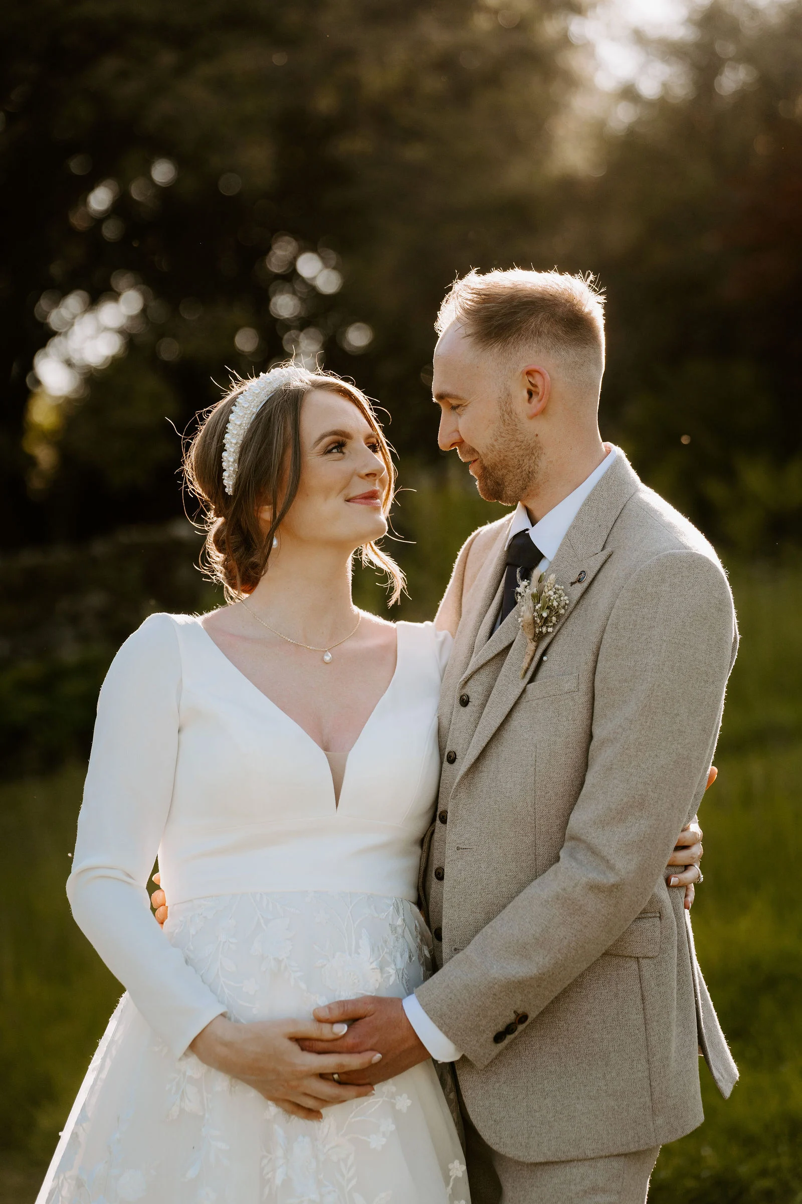 A bride in a long-sleeved white gown and pearl headband and a groom in a tweed suit with a dried-flower buttonhole smile at each other in low golden evening light, with soft green trees behind them.