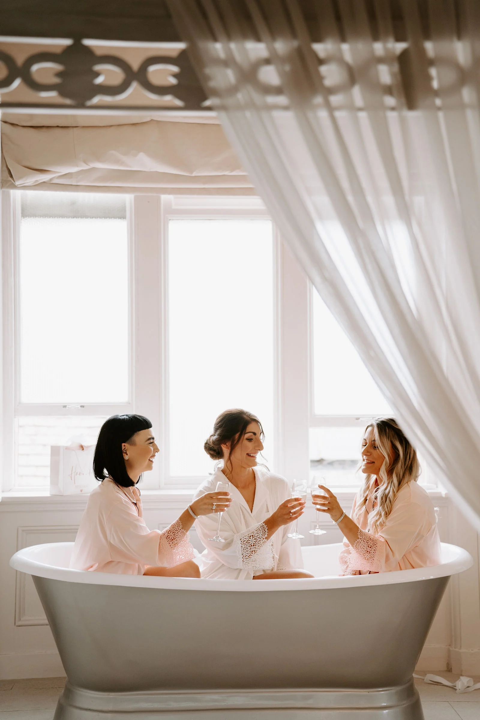 Three women in white robes sit in a freestanding bath in front of a tall window hung with white curtains, raising their glasses