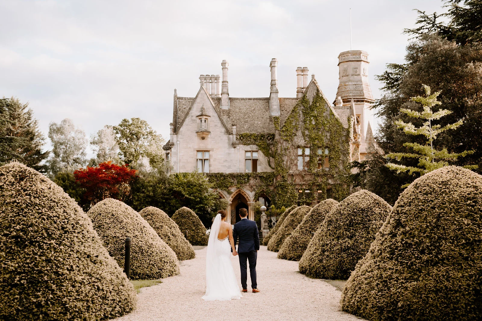 A bride in a long veil and a groom in a navy suit hold hands and walk away from the camera along a gravel path between rounded clipped hedges towards an ivy-covered stone manor house with a tower.