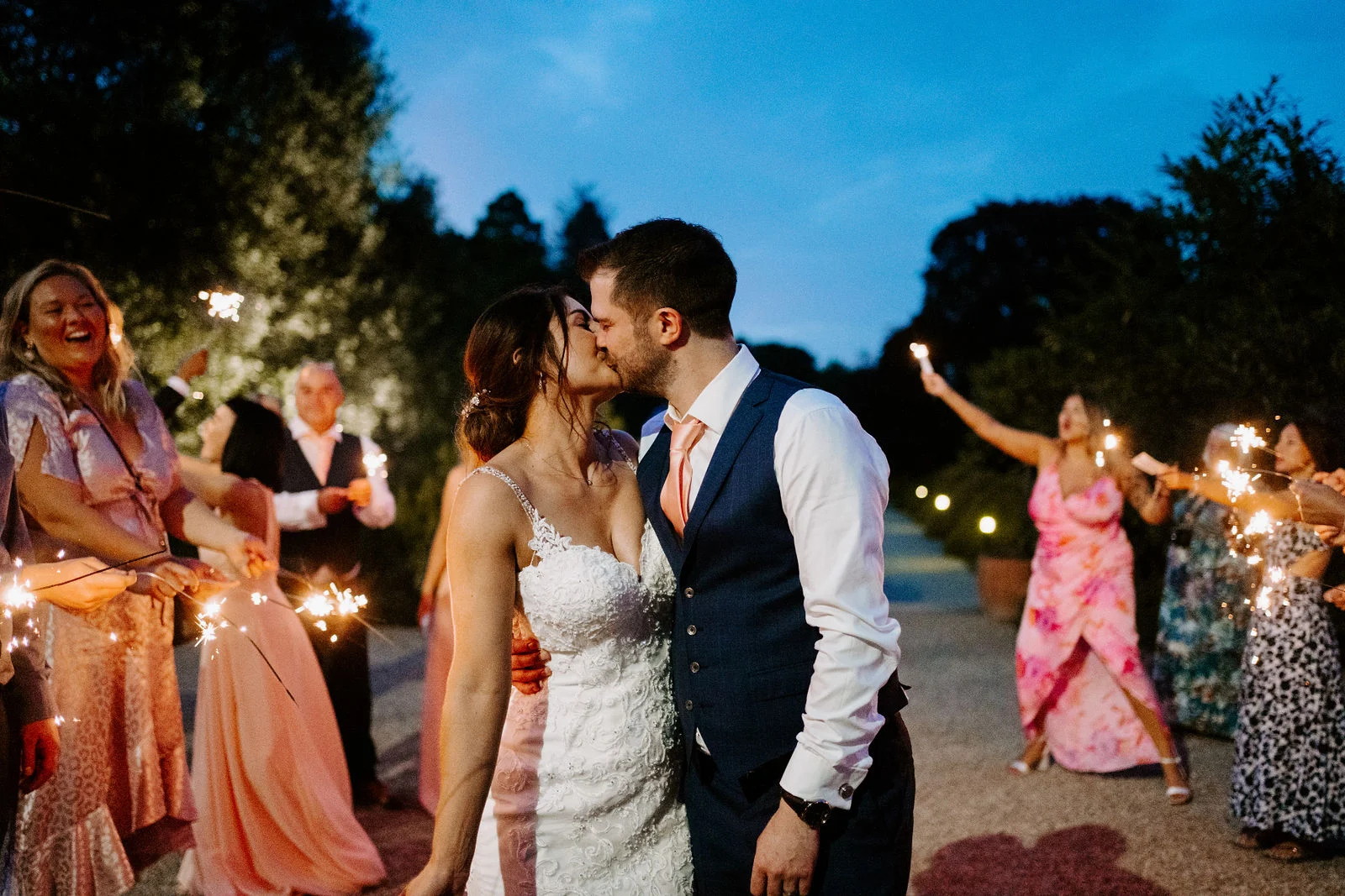 A bride and groom kiss outdoors at dusk while guests in bright dresses hold sparklers around them
