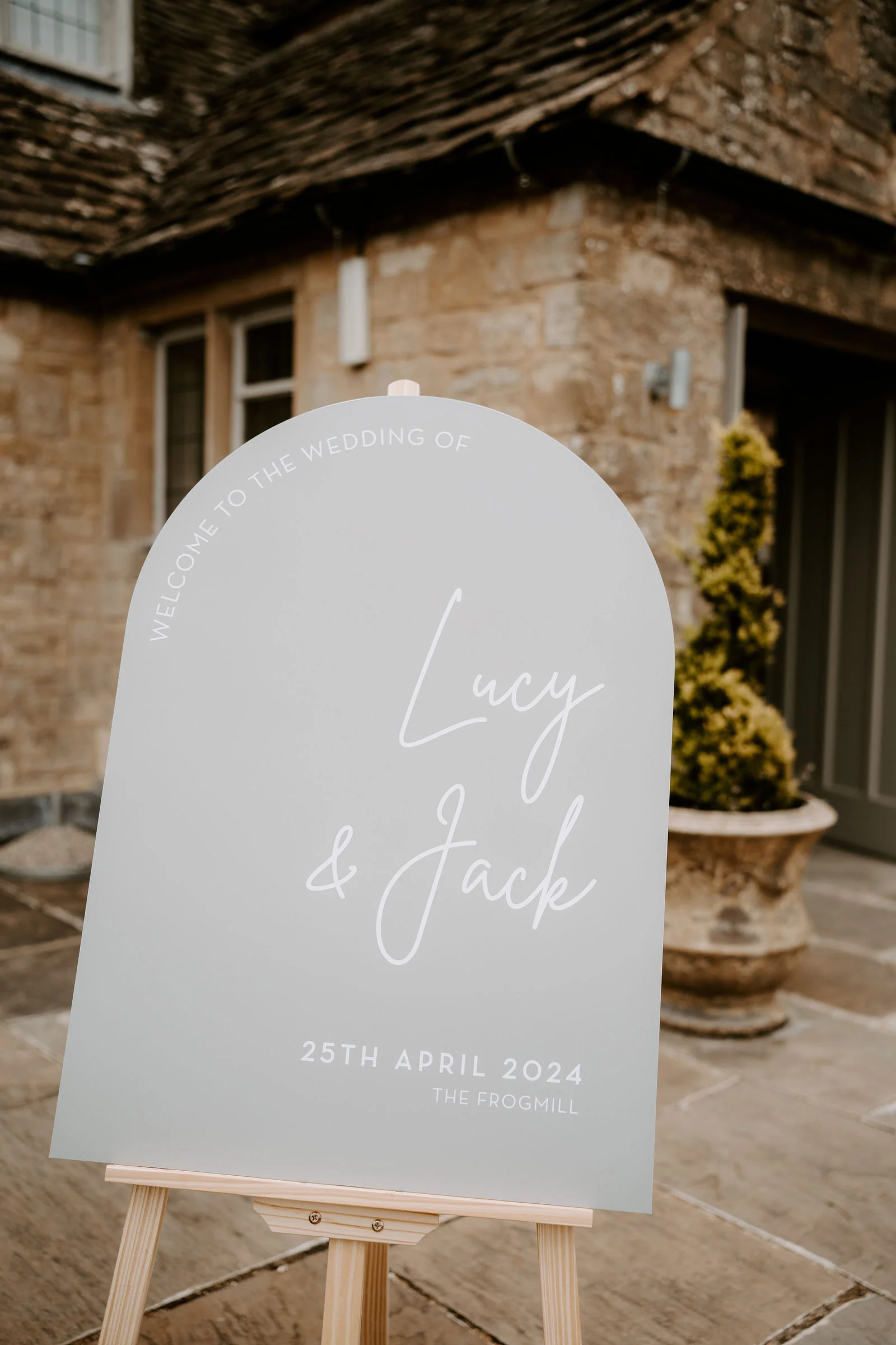 A pale green arched welcome sign on an easel showing the couple’s names and wedding date outside a stone building