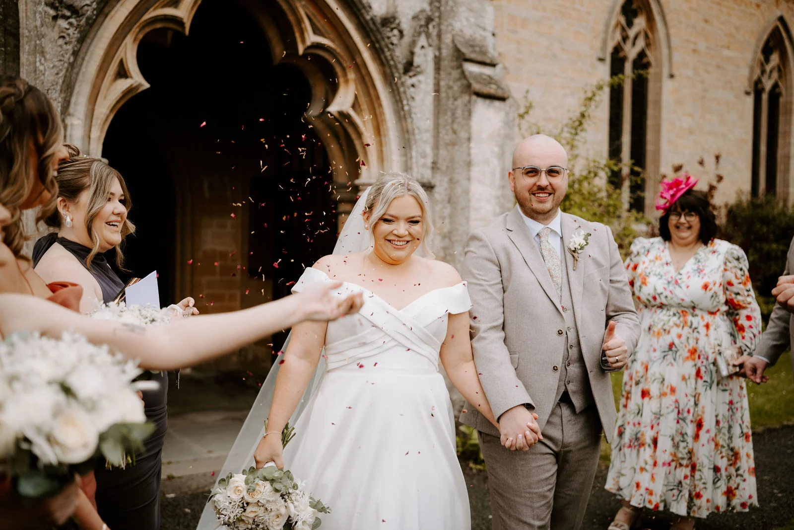 A smiling bride in a white gown and a groom in a light grey suit walk hand in hand out of a pointed stone church porch while petals of confetti fall around them.