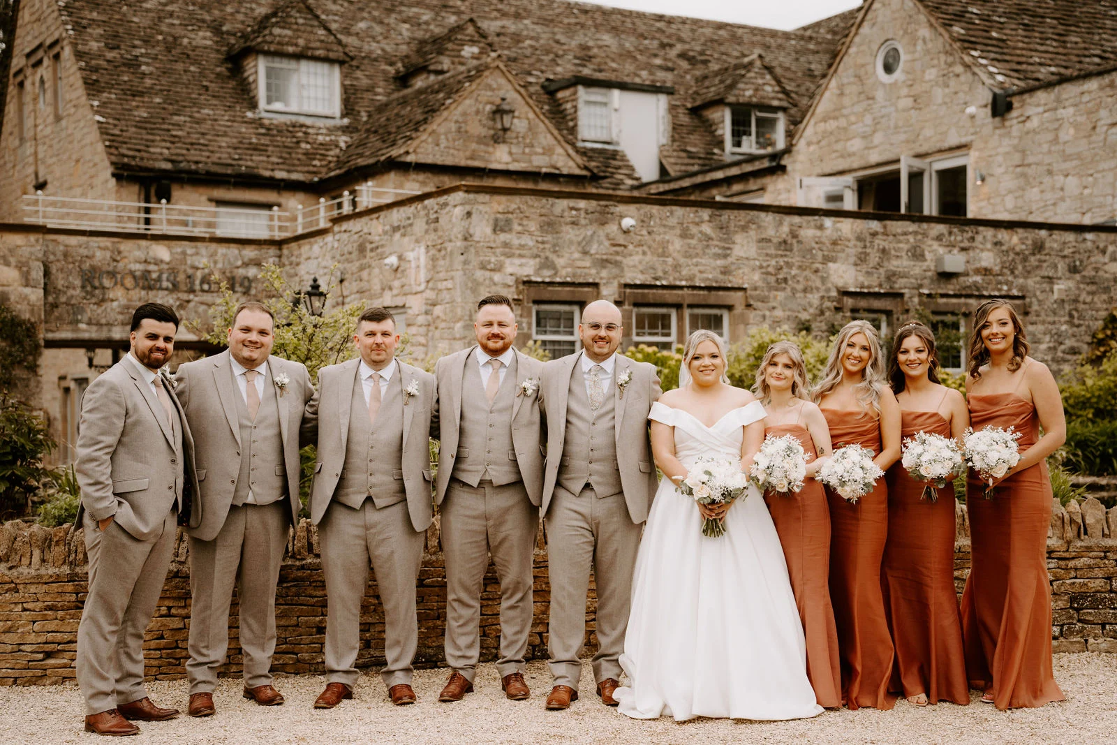 A bride in a white off-the-shoulder gown stands in a row with five men in light grey suits and four bridesmaids in terracotta dresses on gravel in front of a stone inn with a tiled gabled roof.