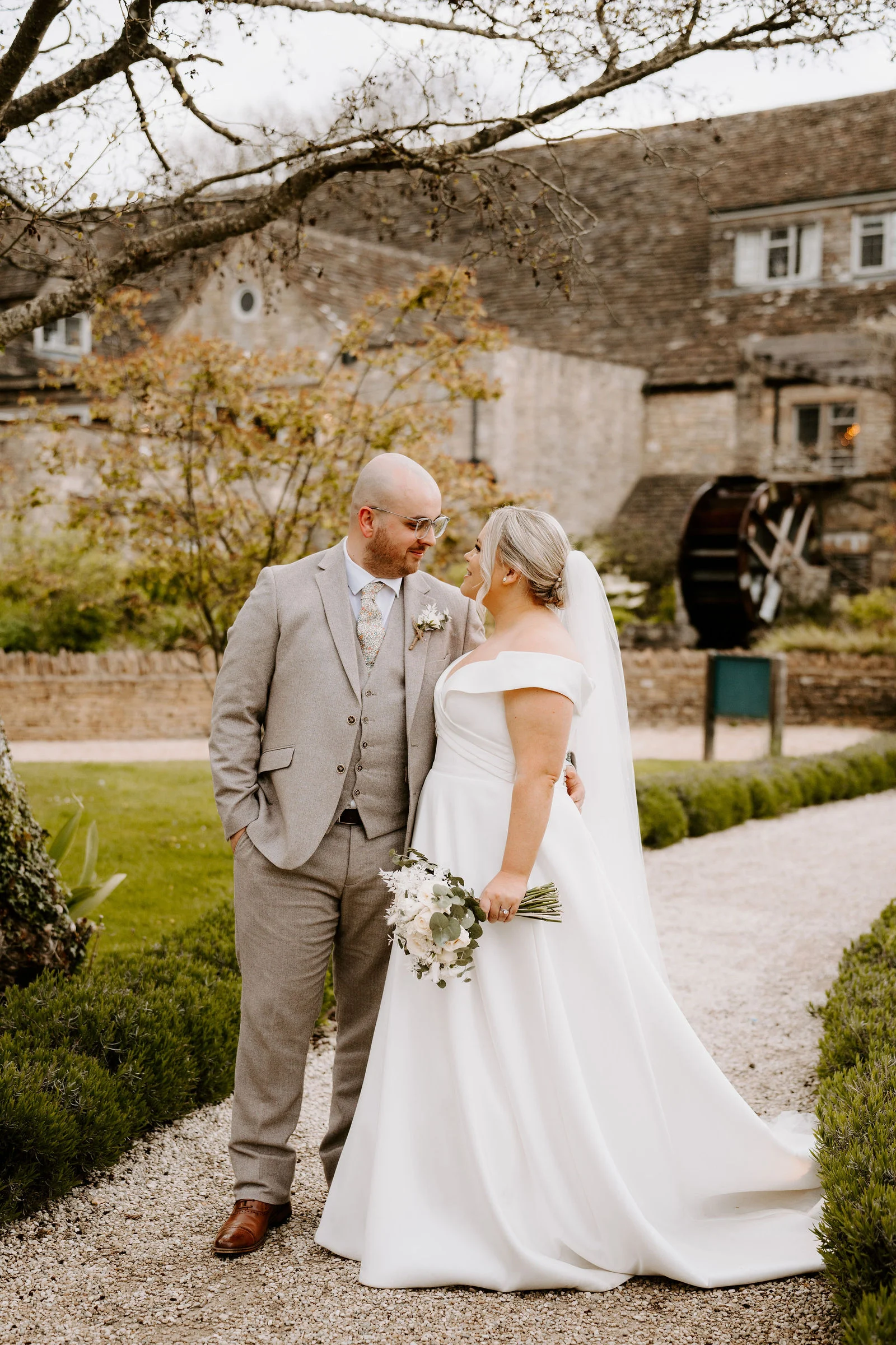 A groom in a light grey three-piece suit and a bride in a white off-the-shoulder gown with a veil and white bouquet smile at each other on a gravel path beside a lawn, with a stone building and a water wheel behind them.