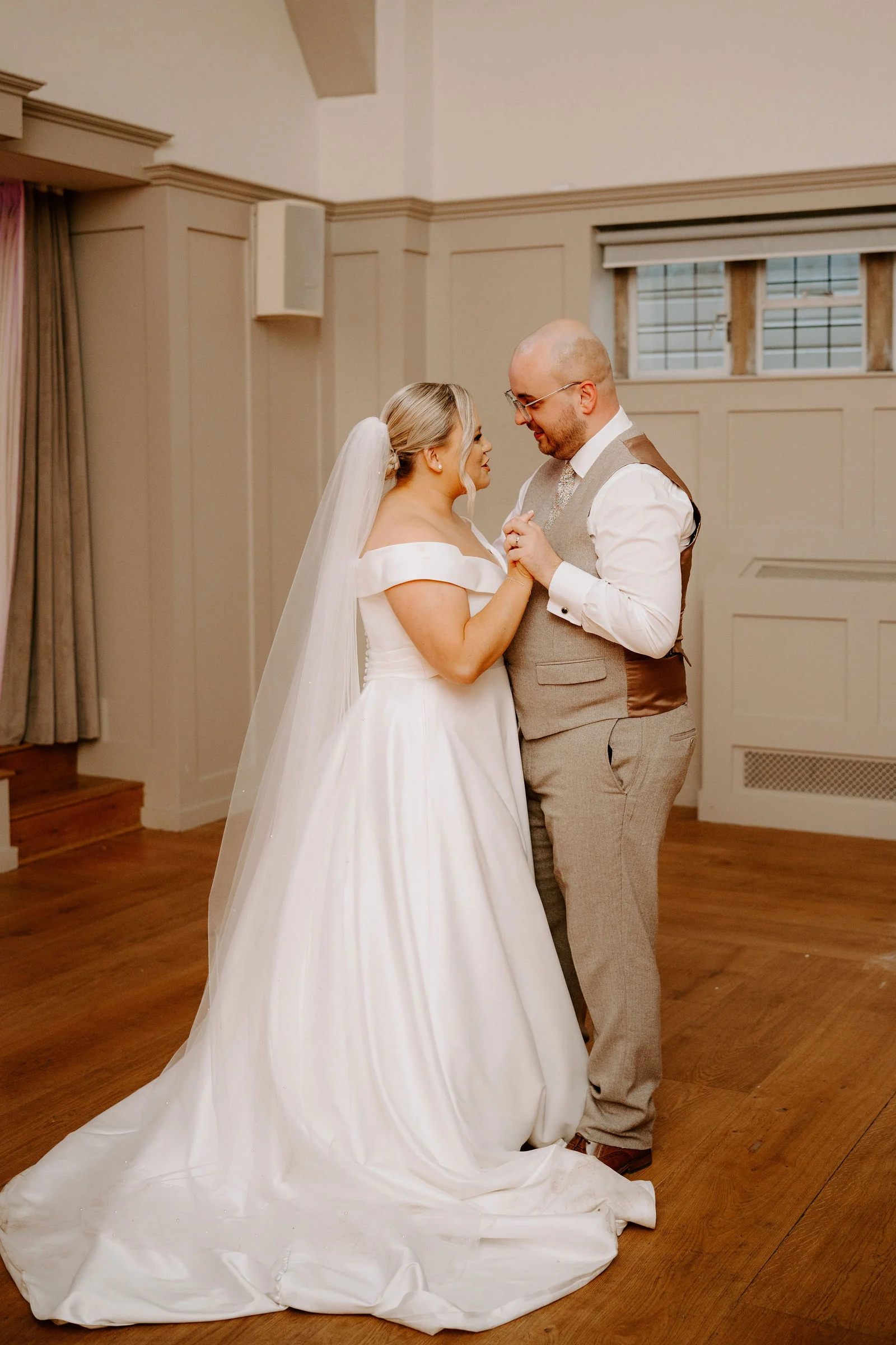 A bride in a white gown with a long veil and a groom in a waistcoat and shirtsleeves hold hands and smile at each other on a wooden dance floor in a panelled room with leaded windows.