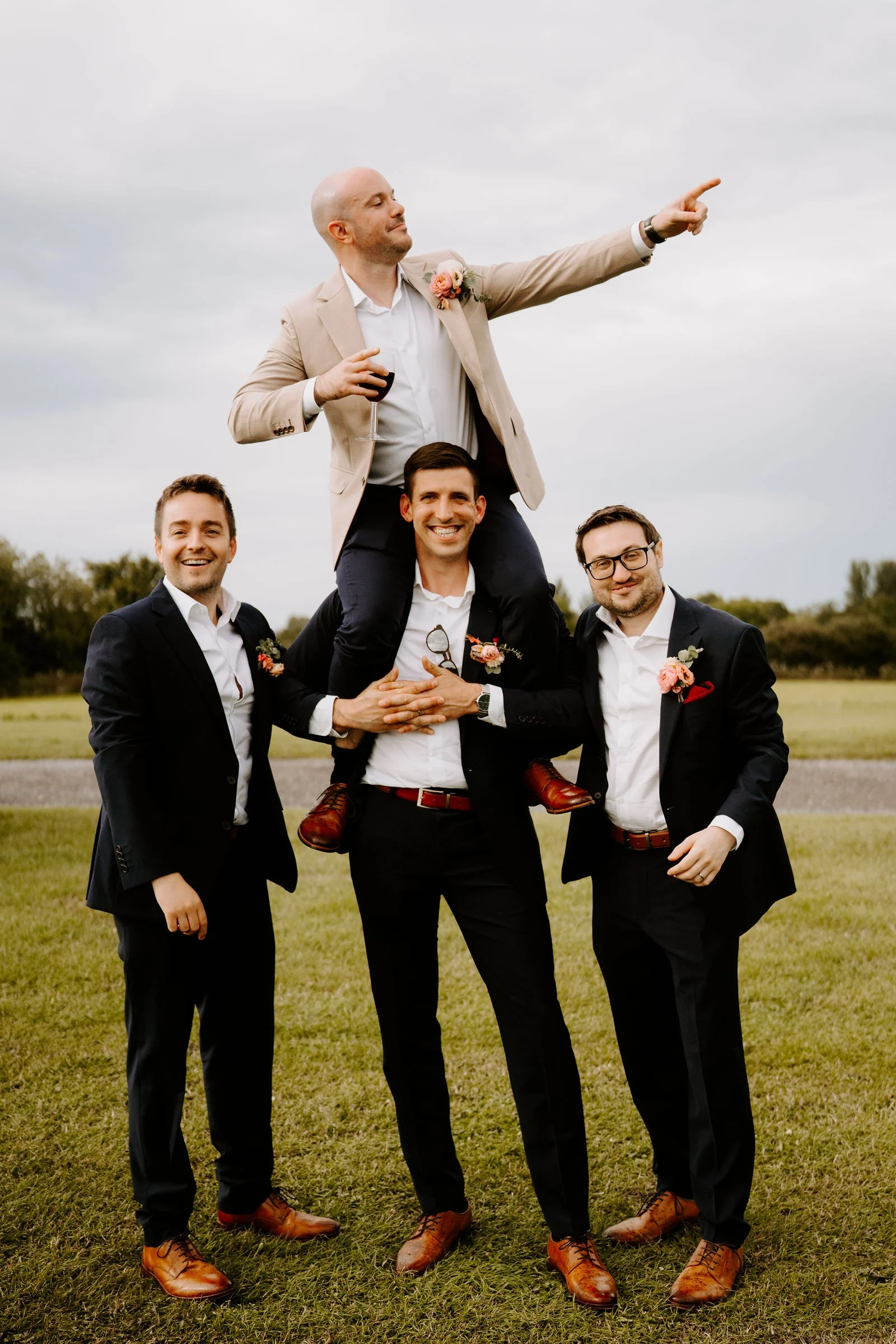 A groom in a beige jacket sits on the shoulders of a groomsman in the middle of two others in dark suits and points into the distance, all laughing on a lawn with trees behind