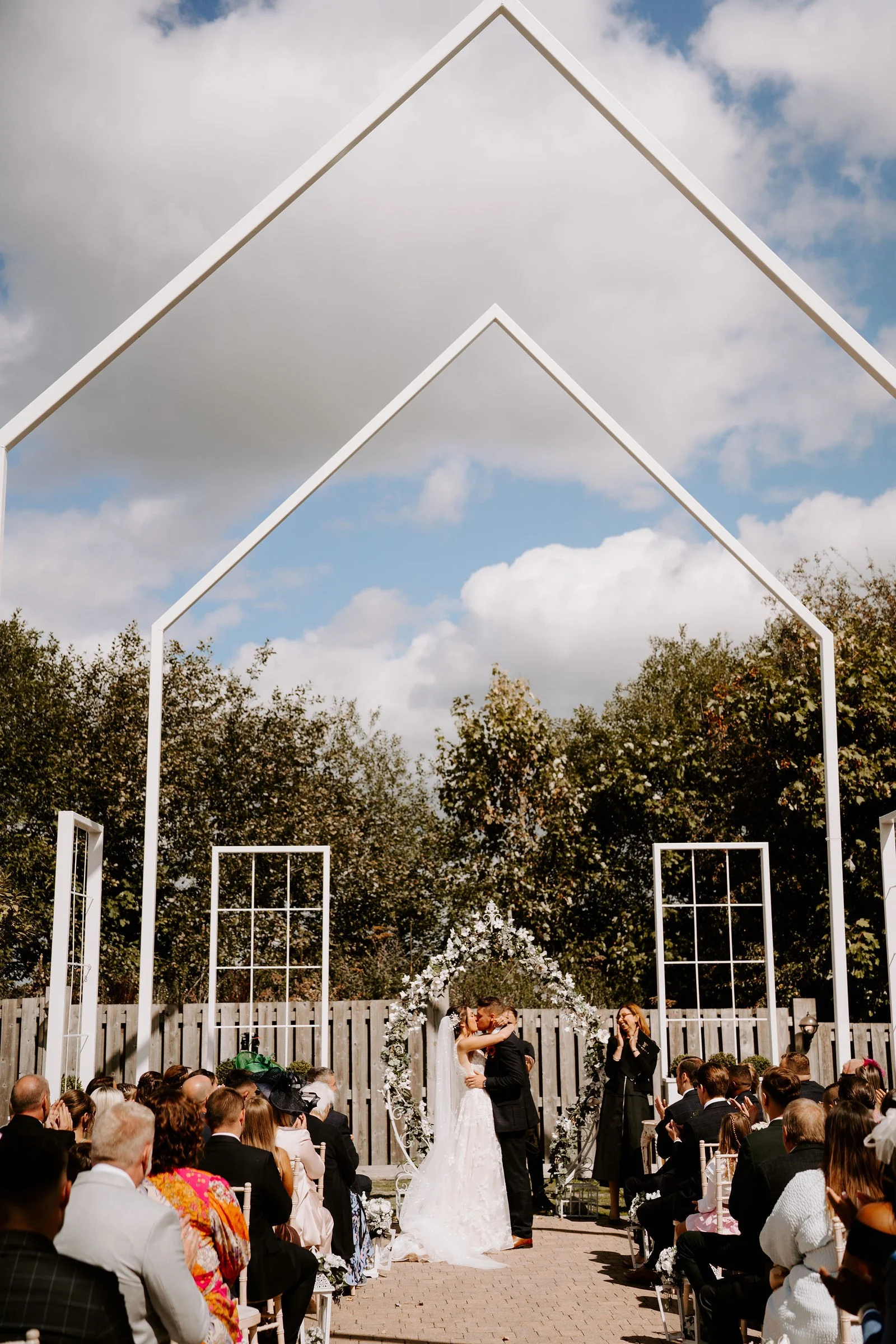 A wedding ceremony seen from behind the seated guests at the White House, the couple kissing beneath a floral arch in front of tall white open frames shaped like a house and window frames, with trees and a timber fence behind