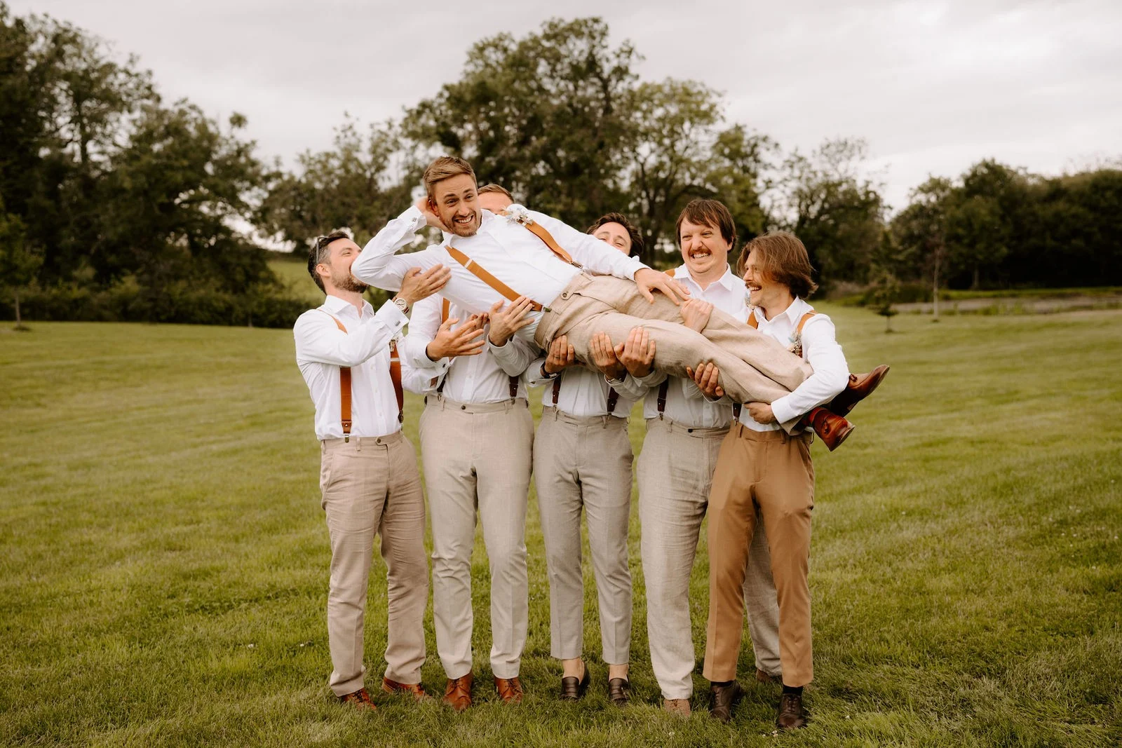 A laughing groom in a white shirt and linen trousers is held flat out in the air by five groomsmen on a lawn with trees behind
