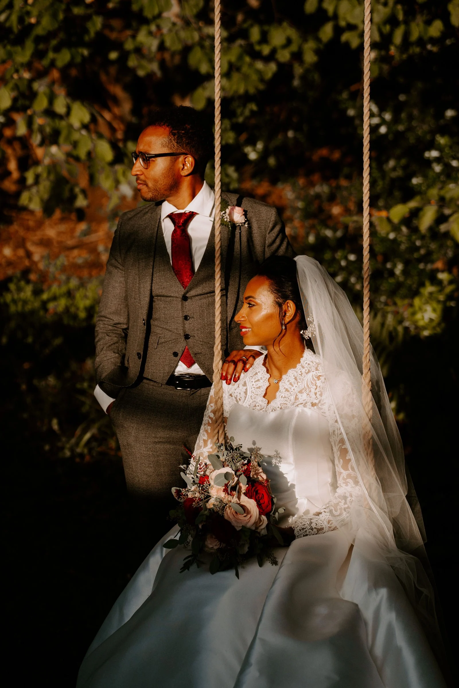 A groom in a grey suit with a red tie and glasses stands with a hand on his bride's shoulder as she sits on a rope swing in a veil, a bouquet in her lap, in dark orange evening light under the trees