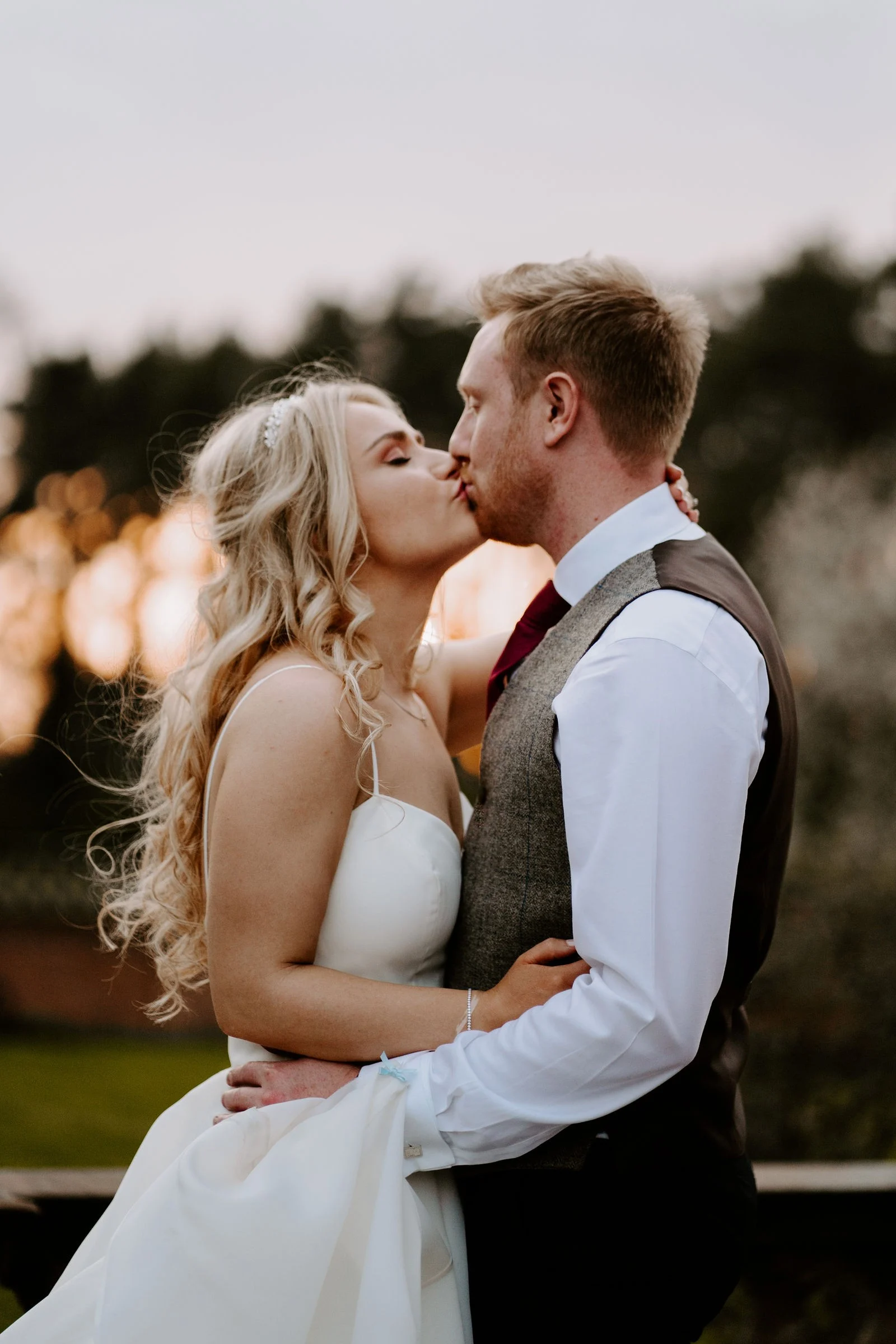 A bride with long curled hair and a groom in a tweed waistcoat kiss outdoors with their arms around each other as the low sun glows orange in the trees behind them at Inglewood Manor
