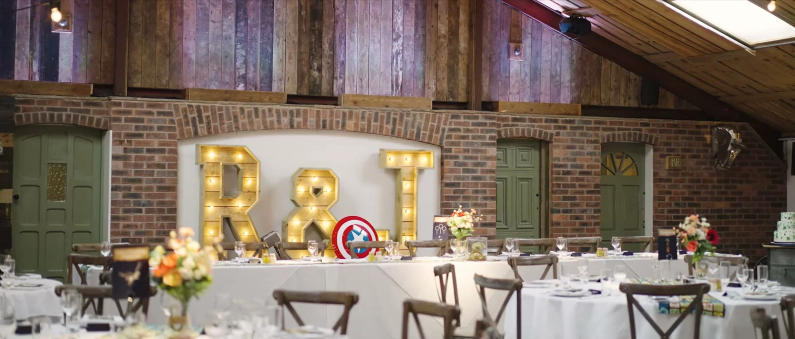 A still from the highlight film: the brick-walled barn at Owen House Wedding Barn set for the meal, with giant lit R and J letters, round tables in white cloths, wooden cross-back chairs and orange and peach flowers