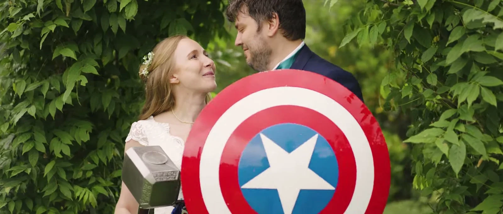 A still from the highlight film: a bride with a flower in her hair looks up at her groom among green leaves, with a Captain America style shield and a toy hammer in front of them at Owen House Wedding Barn