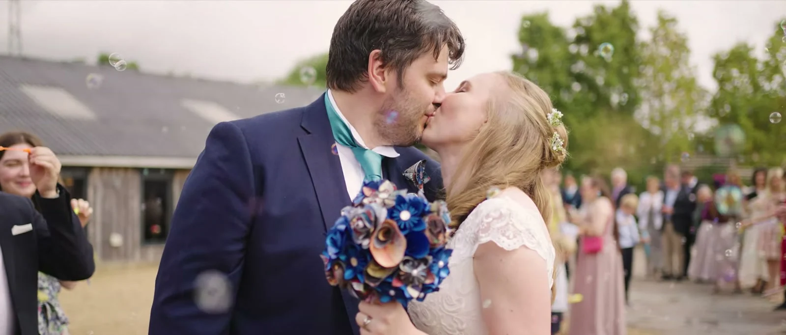 A still from the highlight film: a groom in a navy suit and teal tie kisses his bride, who holds a bouquet of blue and copper flowers, in a shower of bubbles with guests lining the courtyard at Owen House Wedding Barn