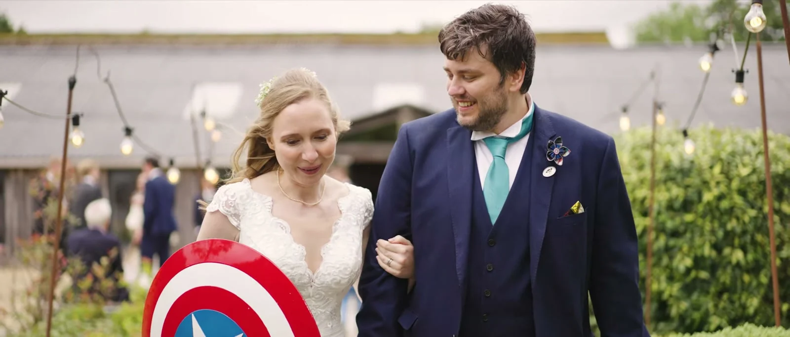 A still from the highlight film: a smiling groom in a navy suit and teal tie walks arm in arm with his bride past strings of festoon lights, a red, white and blue shield at her side, at Owen House Wedding Barn