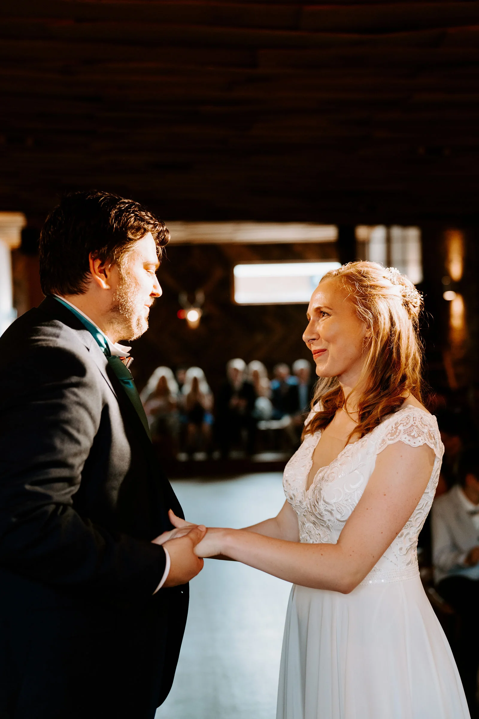 A groom and a bride in a lace dress hold hands facing each other in a dim timbered hall with seated guests blurred behind them