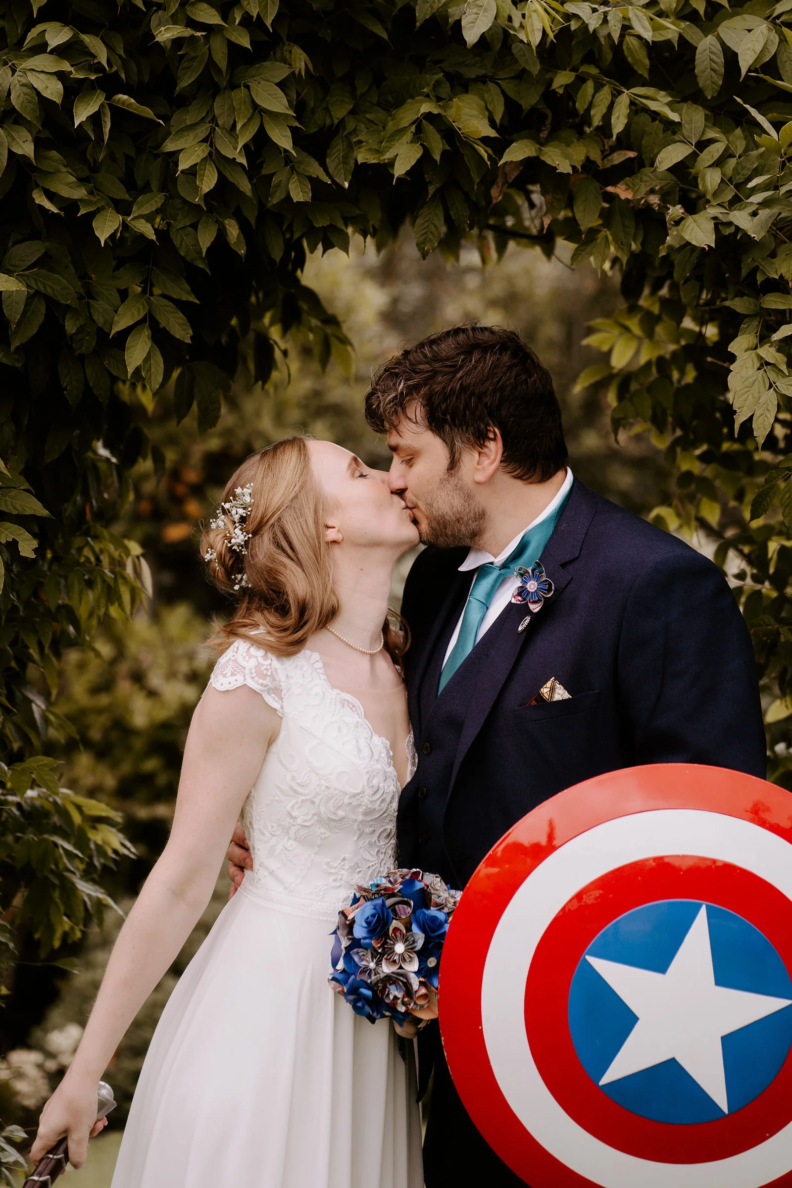 A bride and a groom in a navy suit with a teal tie kiss among green leaves, the groom holding a red, white and blue Captain America shield and the bride a bouquet of blue paper flowers