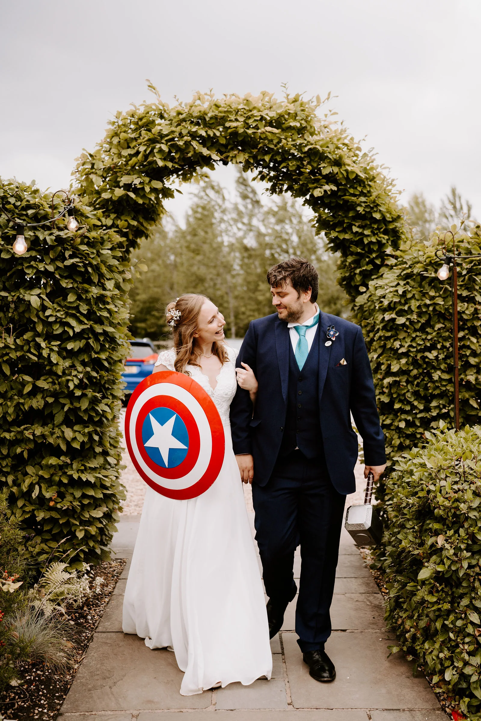 A bride and a groom in a navy suit with a teal tie walk arm in arm along a paved path through a clipped hedge arch strung with festoon lights, the bride carrying a round red, white and blue shield