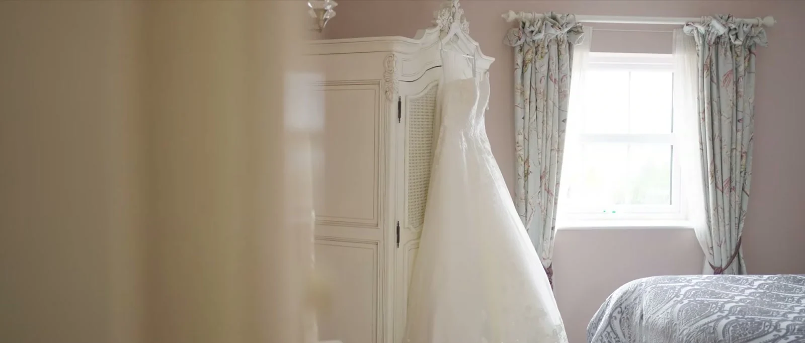 A still from the highlight film: a white wedding dress hangs from a cream wardrobe beside a window with floral curtains in soft light before the wedding at Sandhole Oak Barn