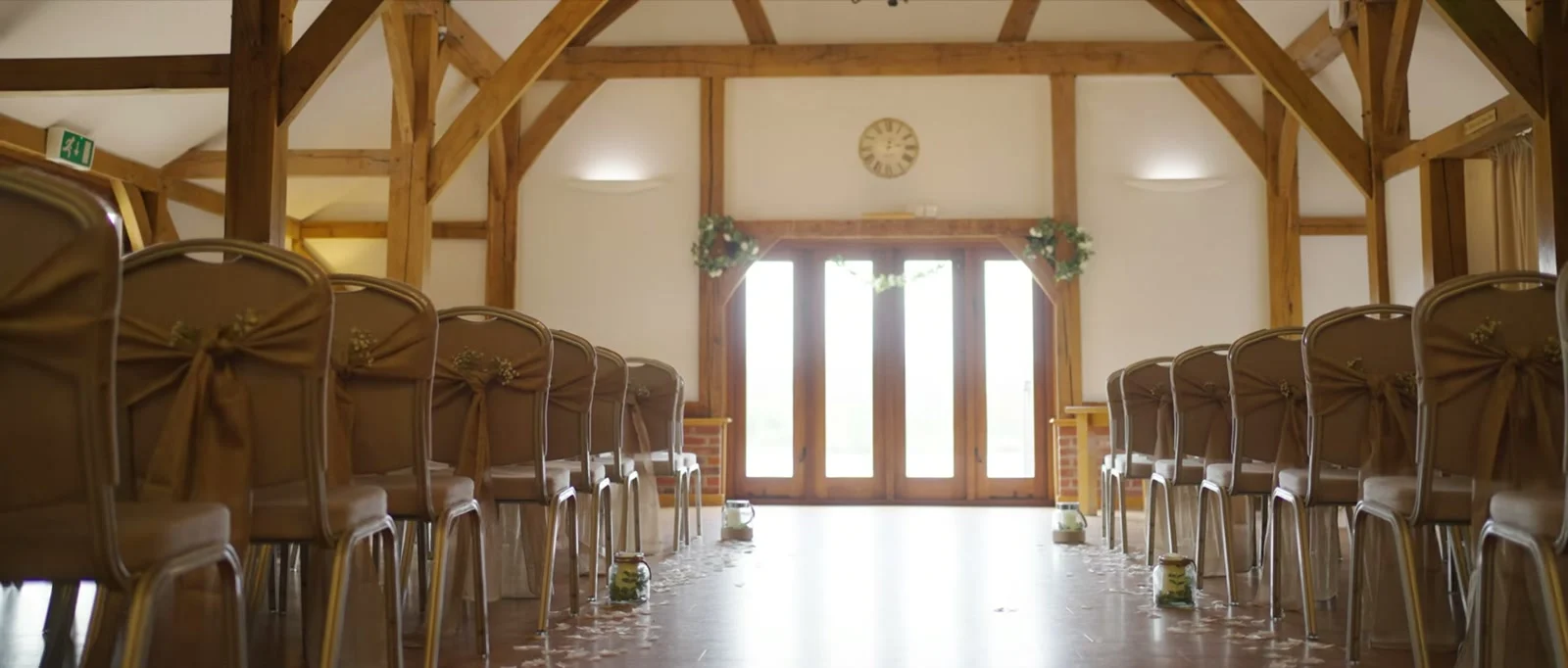 A still from the highlight film: rows of chairs with gold sashes either side of a petal-strewn aisle in an oak-framed barn with tall glazed doors at the far end at Sandhole Oak Barn