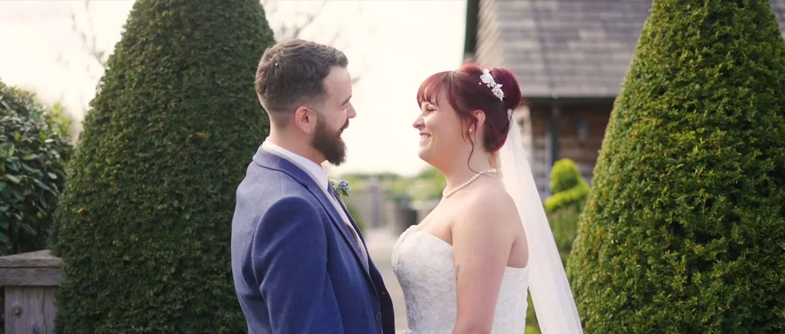 A still from the highlight film: a bearded groom in a blue jacket and a bride in a veil laugh at each other between two clipped yew shapes with a timber-clad building behind at Sandhole Oak Barn