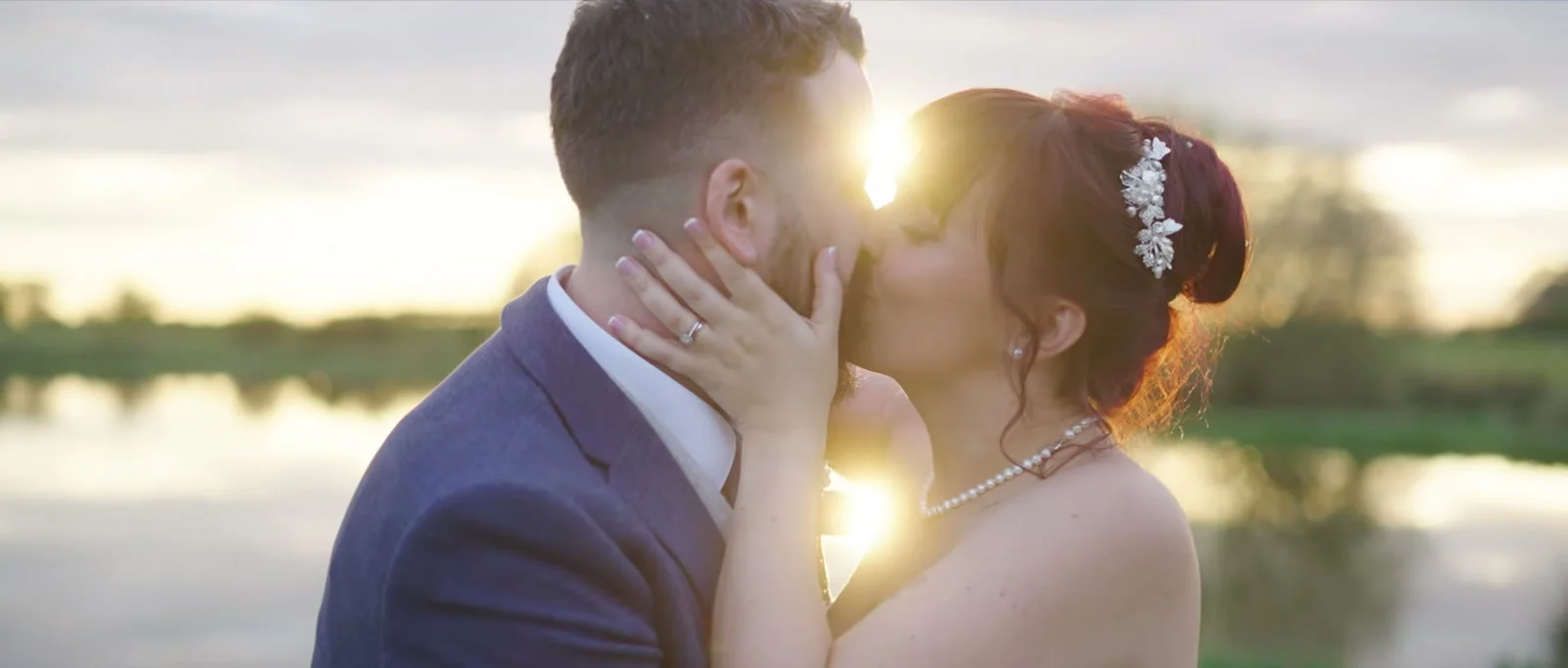 A still from the highlight film: a groom and bride kiss close together by a lake, her hand on his cheek, with the low sun flaring between them at Sandhole Oak Barn
