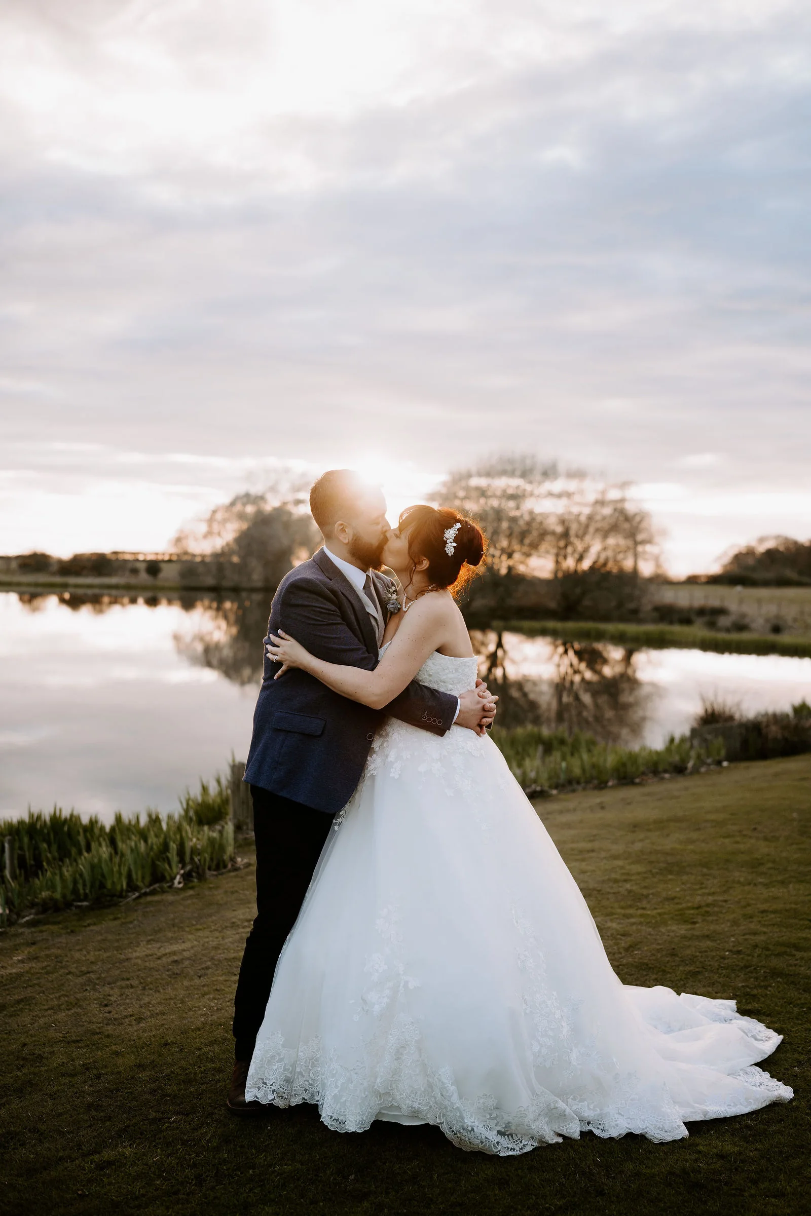 A bride in a full tulle gown and a groom in a blue jacket embrace and kiss beside a still lake, with the low sun behind them