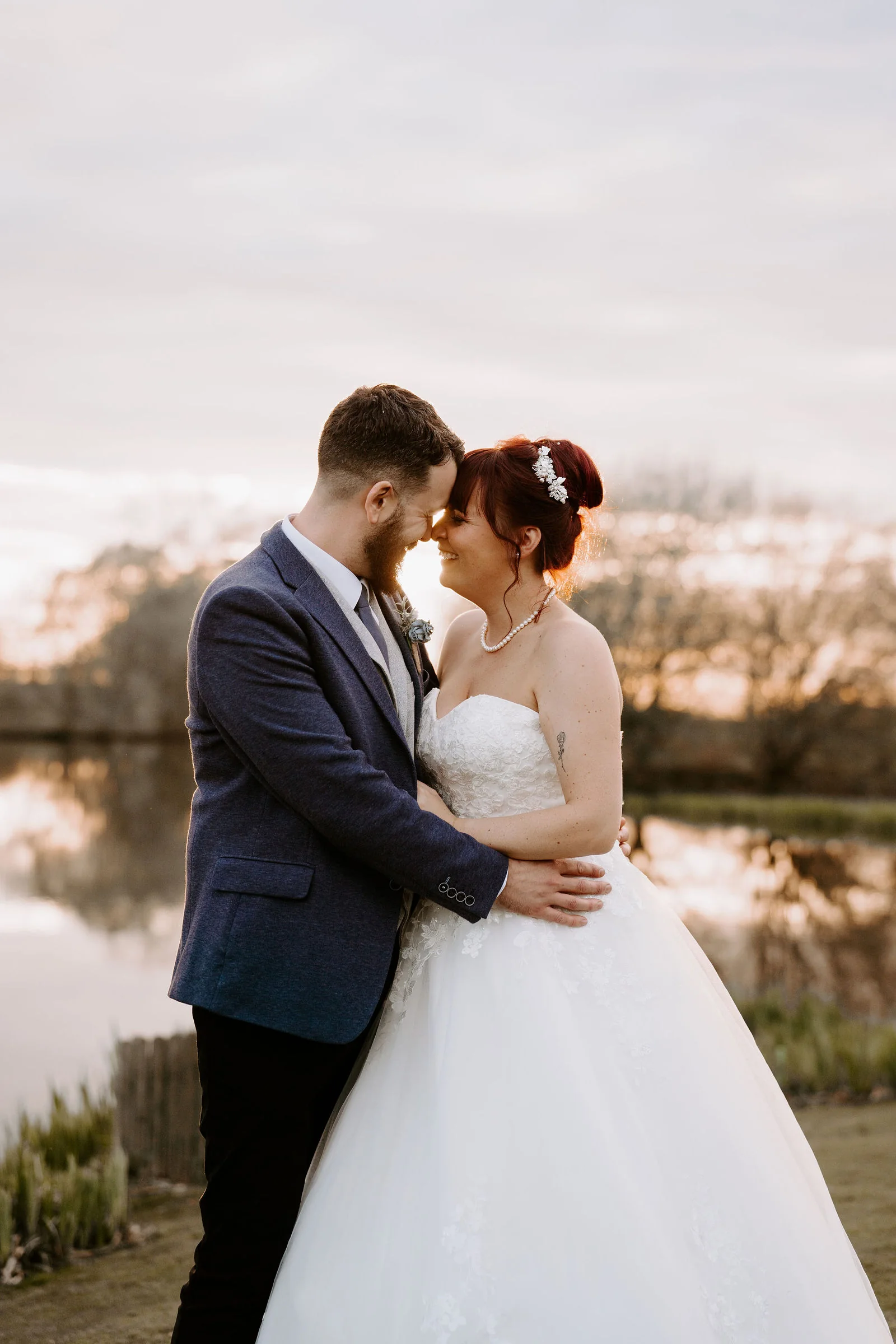 A groom and a bride in a strapless lace gown with a pearl necklace rest their foreheads together beside a lake under a pink dusk sky