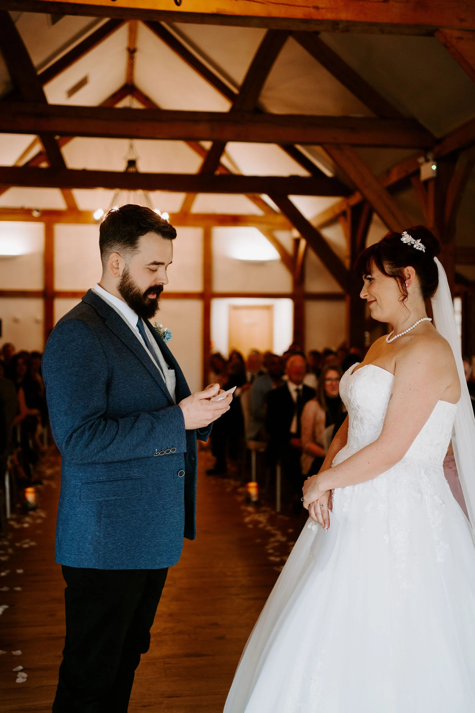A groom in a blue textured jacket looks down at what he holds as he faces his bride in a strapless gown and veil in a timber-framed hall, with guests seated along the aisle