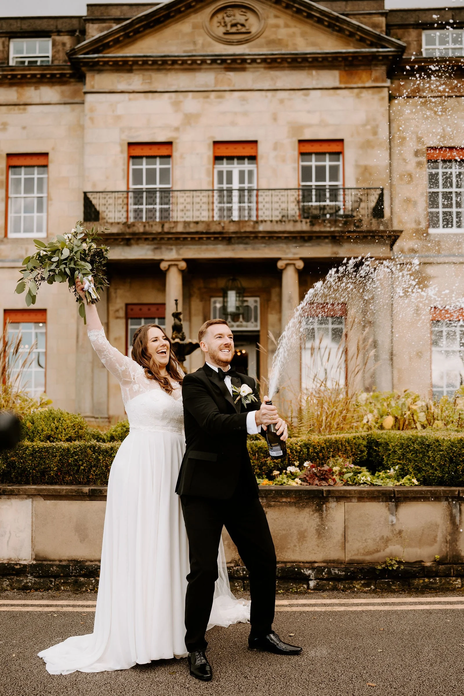 A groom in a dinner suit sprays a bottle of champagne in a wide arc while his laughing bride raises her bouquet, in front of the stone hall with its balcony and columns